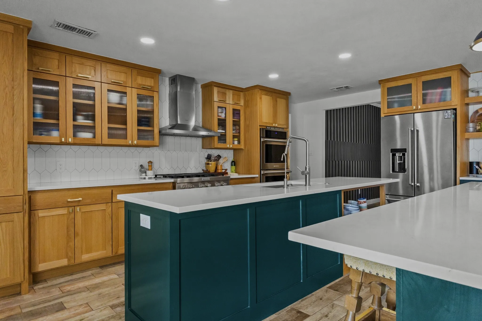 Spacious kitchen featuring light wood-finish cabinetry, a large teal island, white countertops, built-in stainless steel appliances, and wood-finish flooring
