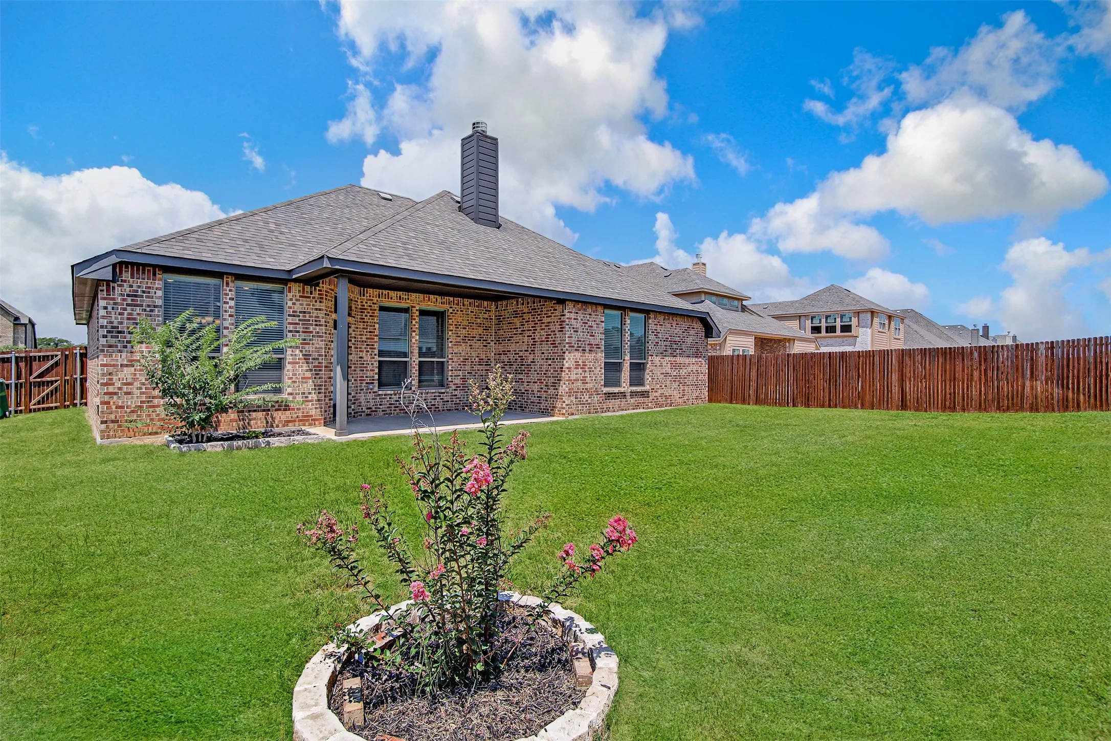 Expansive backyard featuring a manicured lawn, a full brick exterior, a covered patio, a dark composite shingle roof, and a wood privacy fence