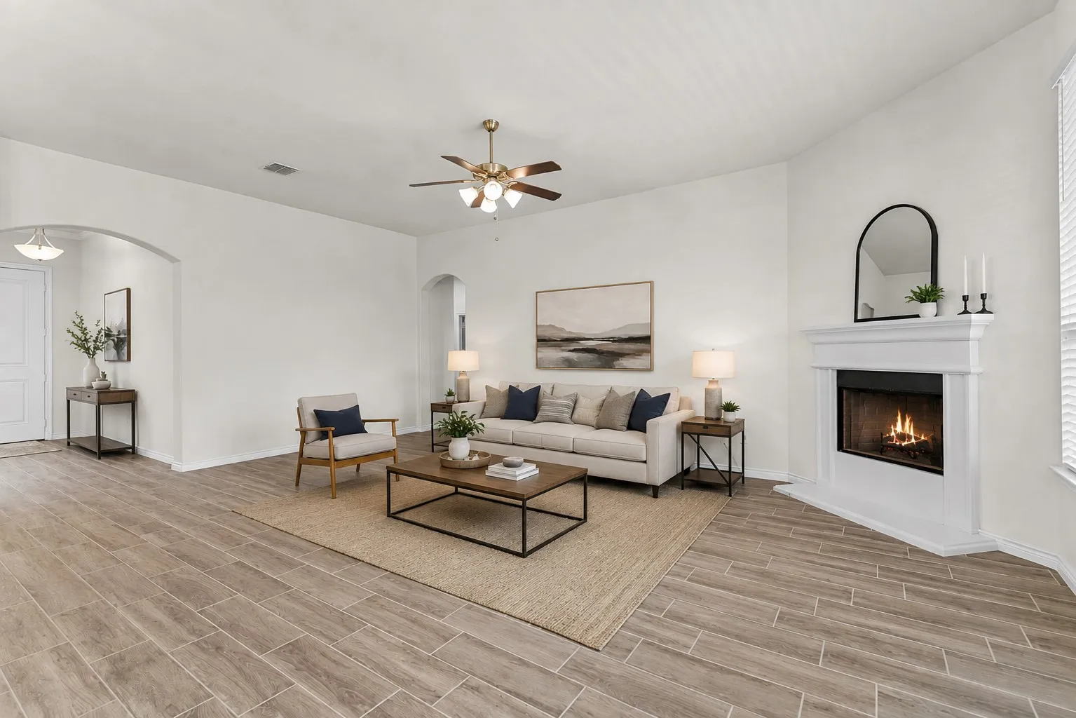 Spacious living room featuring a gas fireplace with a white surround and arched mirror, ceiling fan with integrated lighting, wood-finish plank flooring laid in a herringbone pattern, and a neutral color palette with recessed lighting throughout