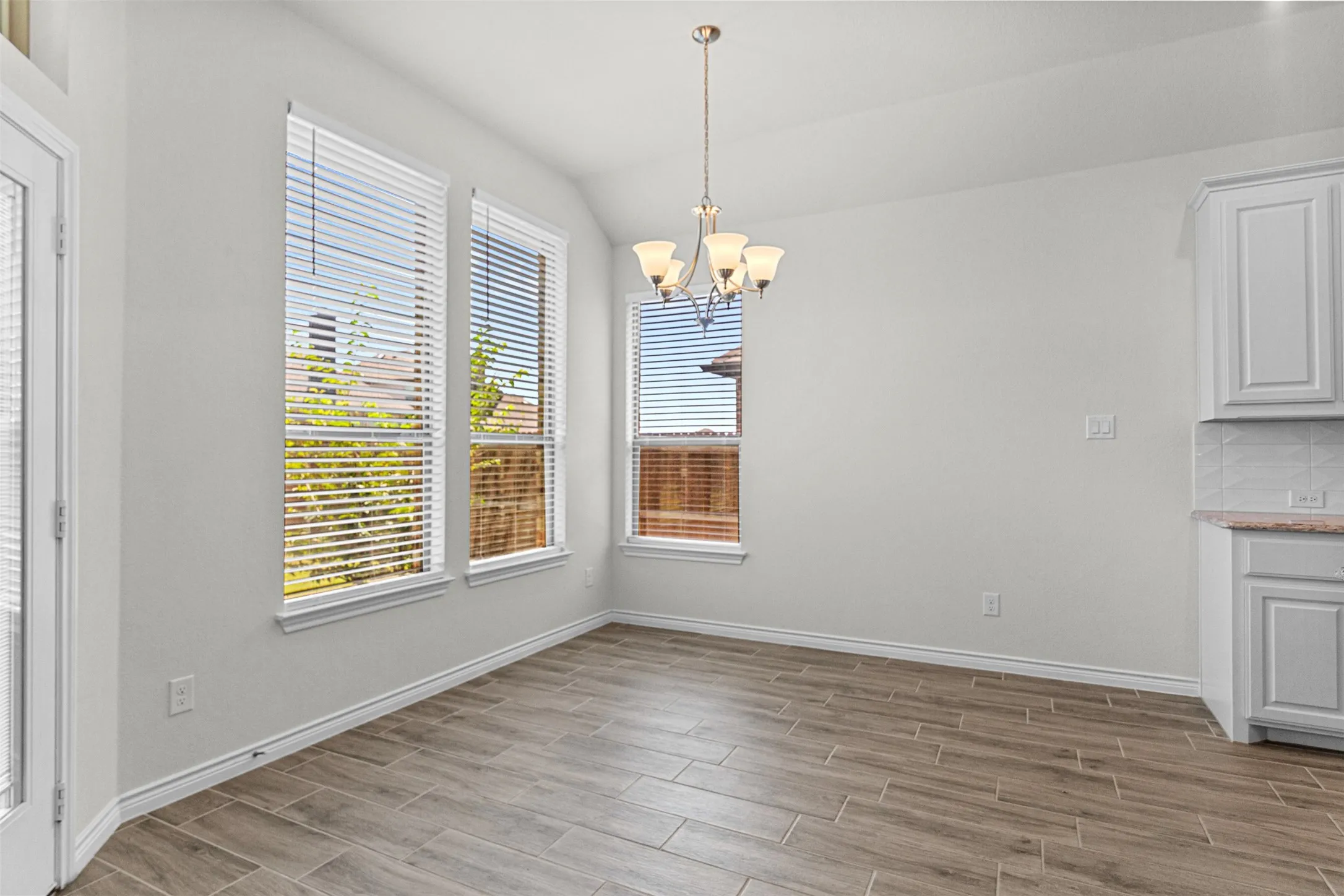 Spacious dining area featuring wood-finish tile flooring, multiple large windows with horizontal blinds, a traditional chandelier, and light-toned cabinetry with a tile backsplash