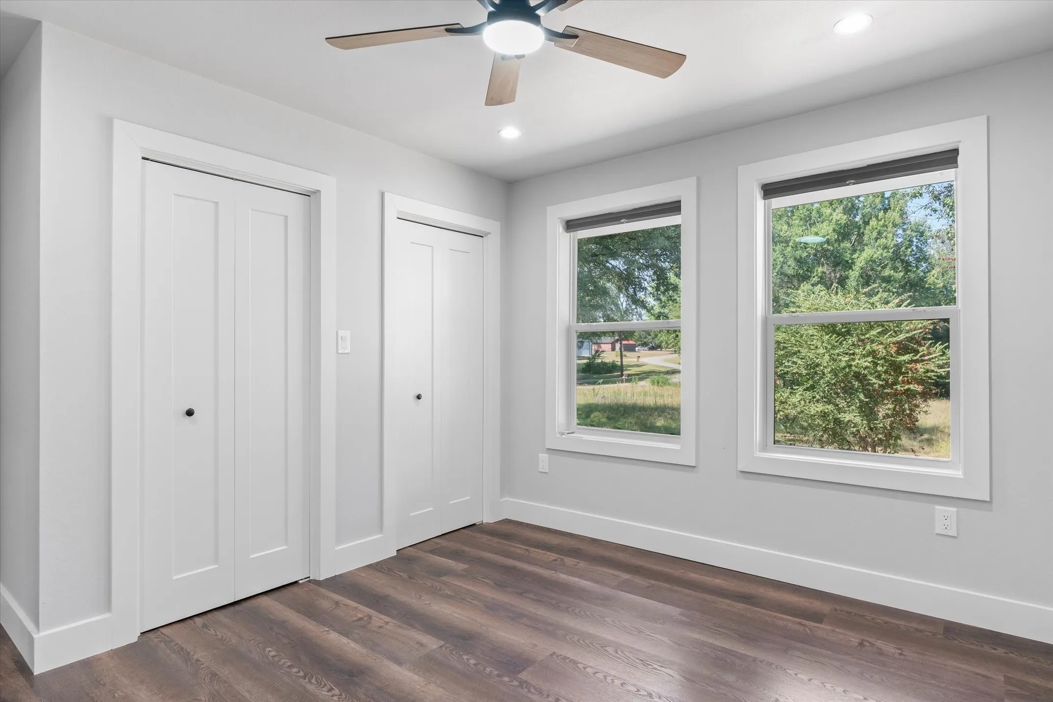 Room featuring wood-finish flooring, two white bi-fold closet doors, two windows with white trim, a ceiling fan with light fixture, and recessed lighting