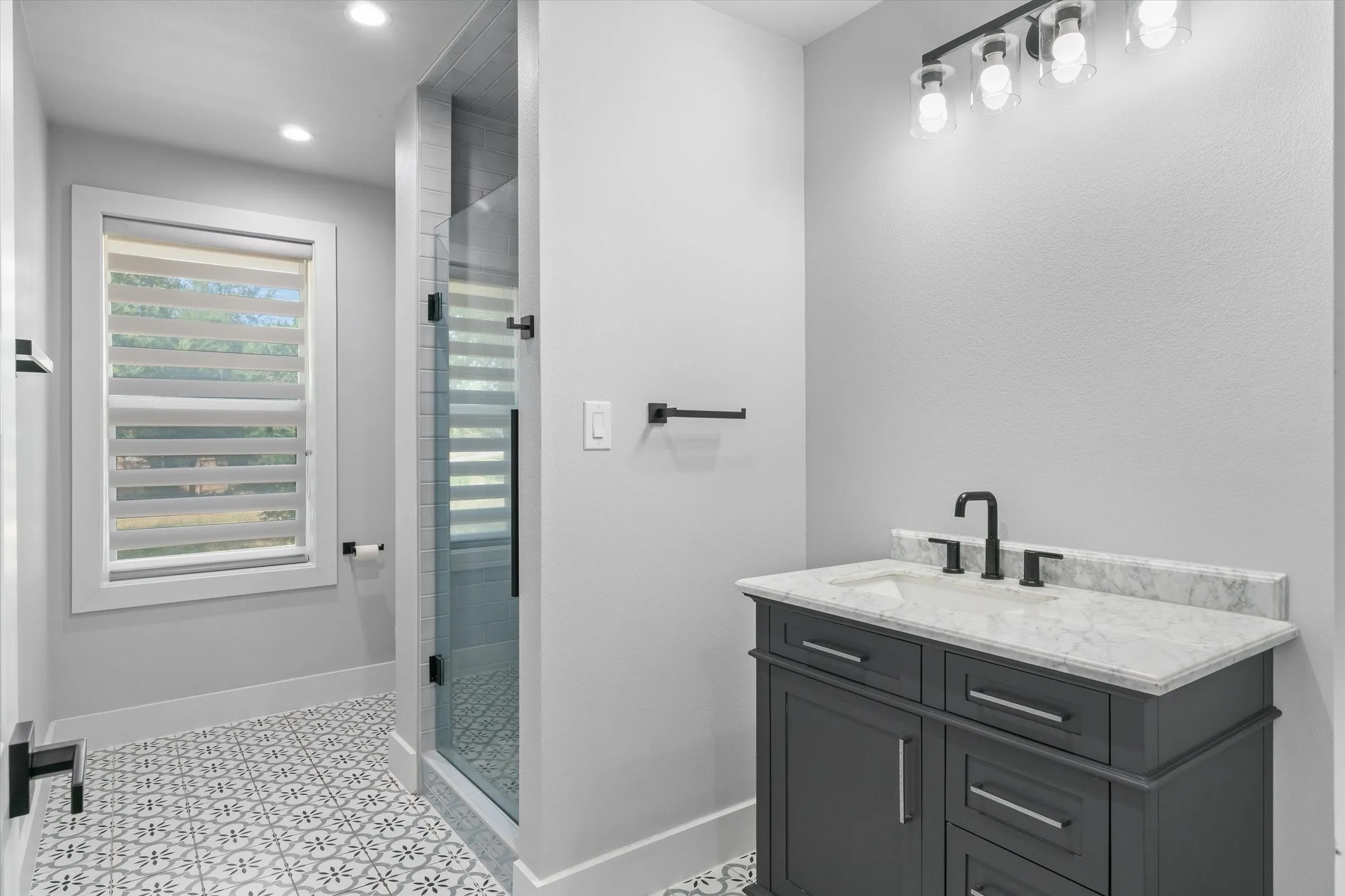 Bathroom featuring a dark gray vanity with a light countertop, a glass-enclosed shower, patterned floor tile, recessed lighting, and a window with blinds