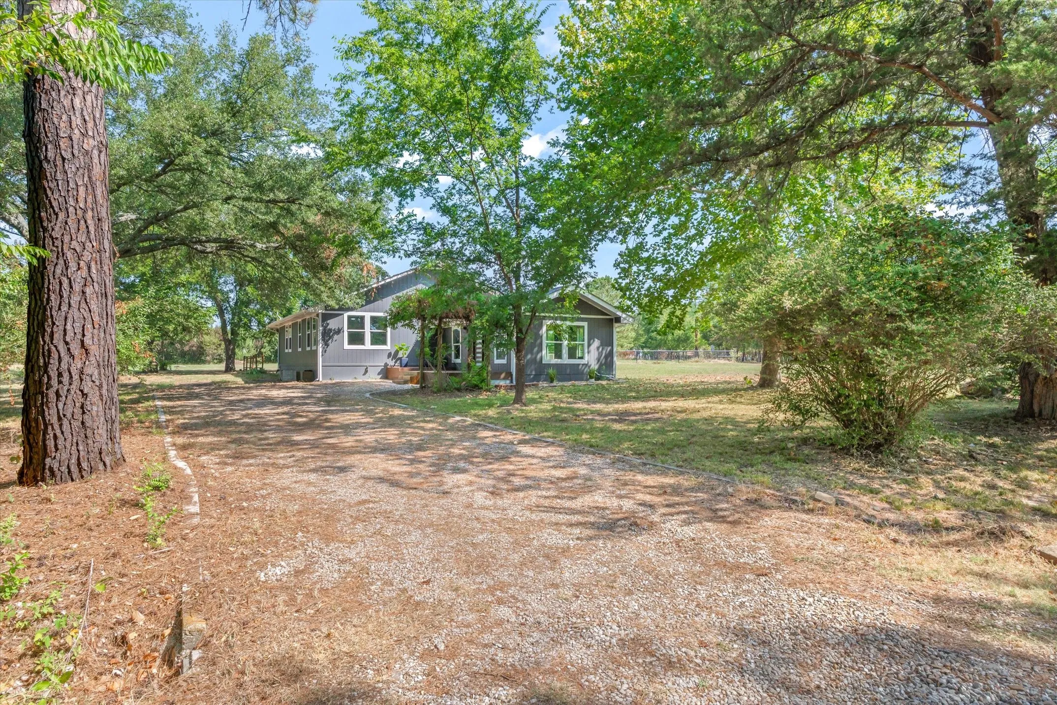 Spacious gravel driveway leading to a gray exterior residence with white-framed windows, surrounded by mature trees and natural landscaping