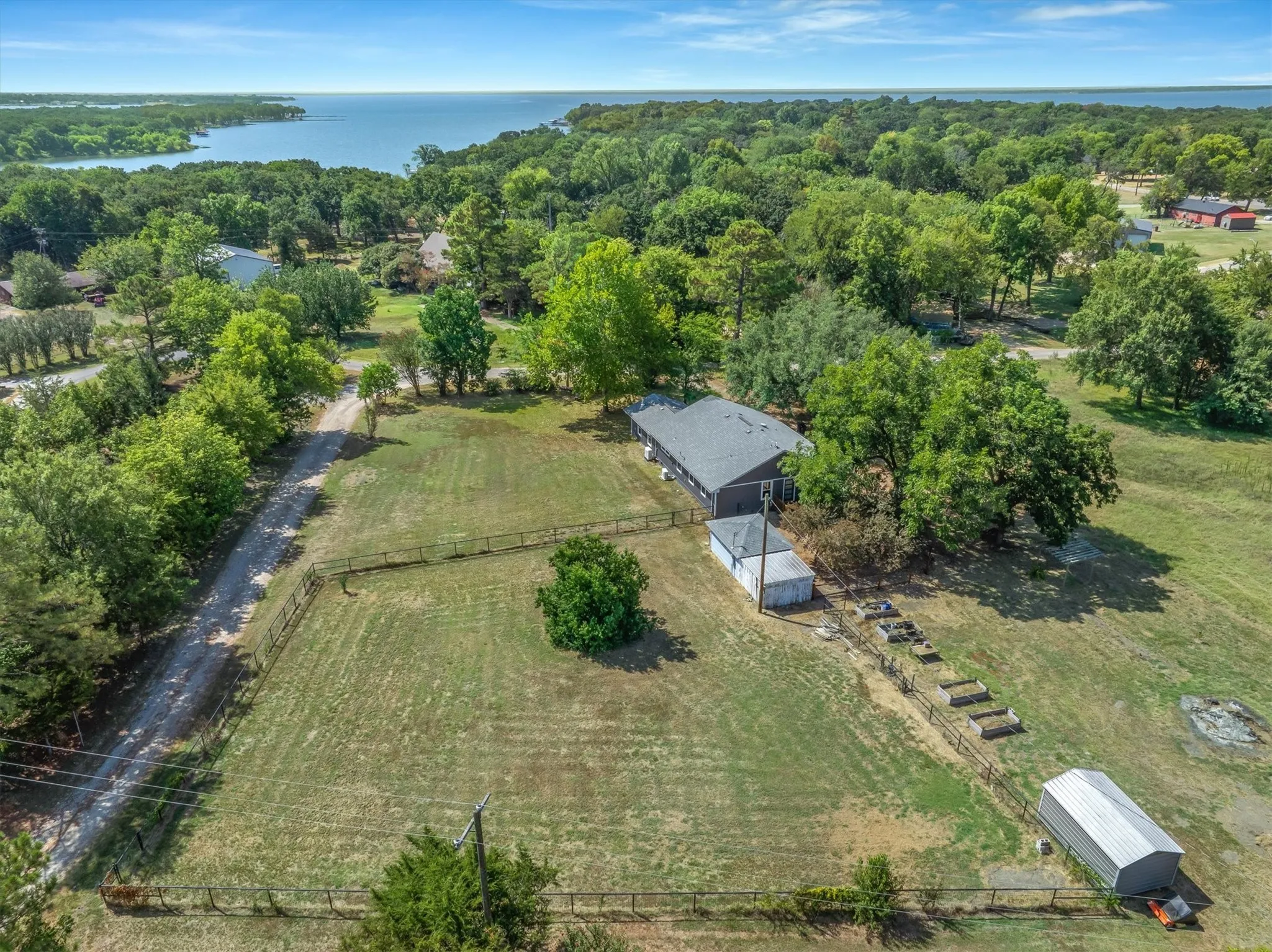 Expansive property featuring a single-story residence with a gray roof, surrounded by mature trees, a fenced yard, and a large body of water in the background