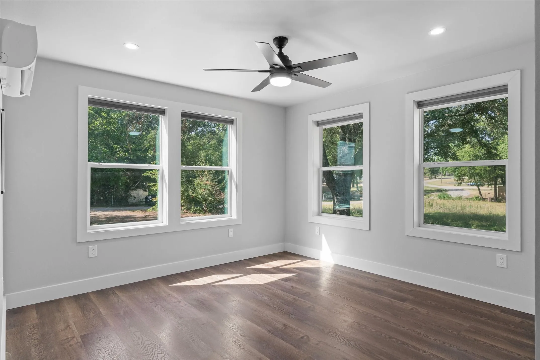 Room featuring five windows with white trim, wood-finish flooring, a ceiling fan with integrated lighting, recessed lighting, and a wall-mounted climate control unit