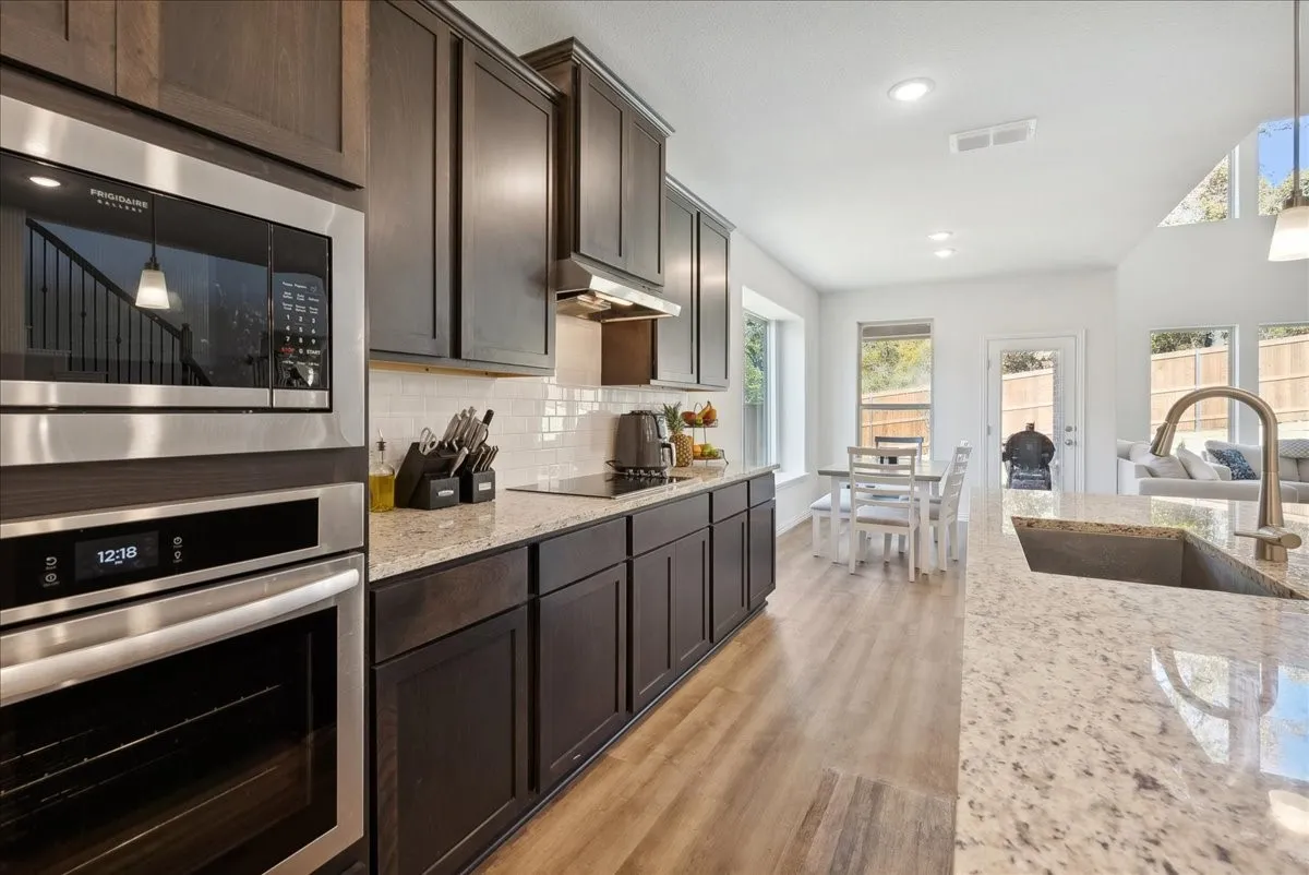 Kitchen featuring dark wood cabinetry, light-toned countertops, subway tile backsplash, stainless steel appliances including a built-in microwave and oven, and wood-finish flooring
