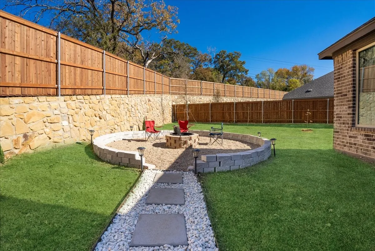 Expansive backyard featuring a built-in circular fire pit area with gravel ground cover, a stone retaining wall, and a wood privacy fence