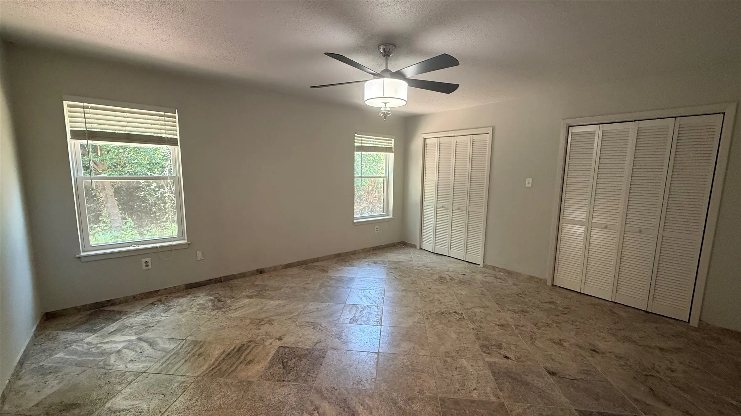 Spacious room featuring large format tile flooring, two windows with blinds, a ceiling fan with integrated lighting, and two sets of bi-fold louvered closet doors