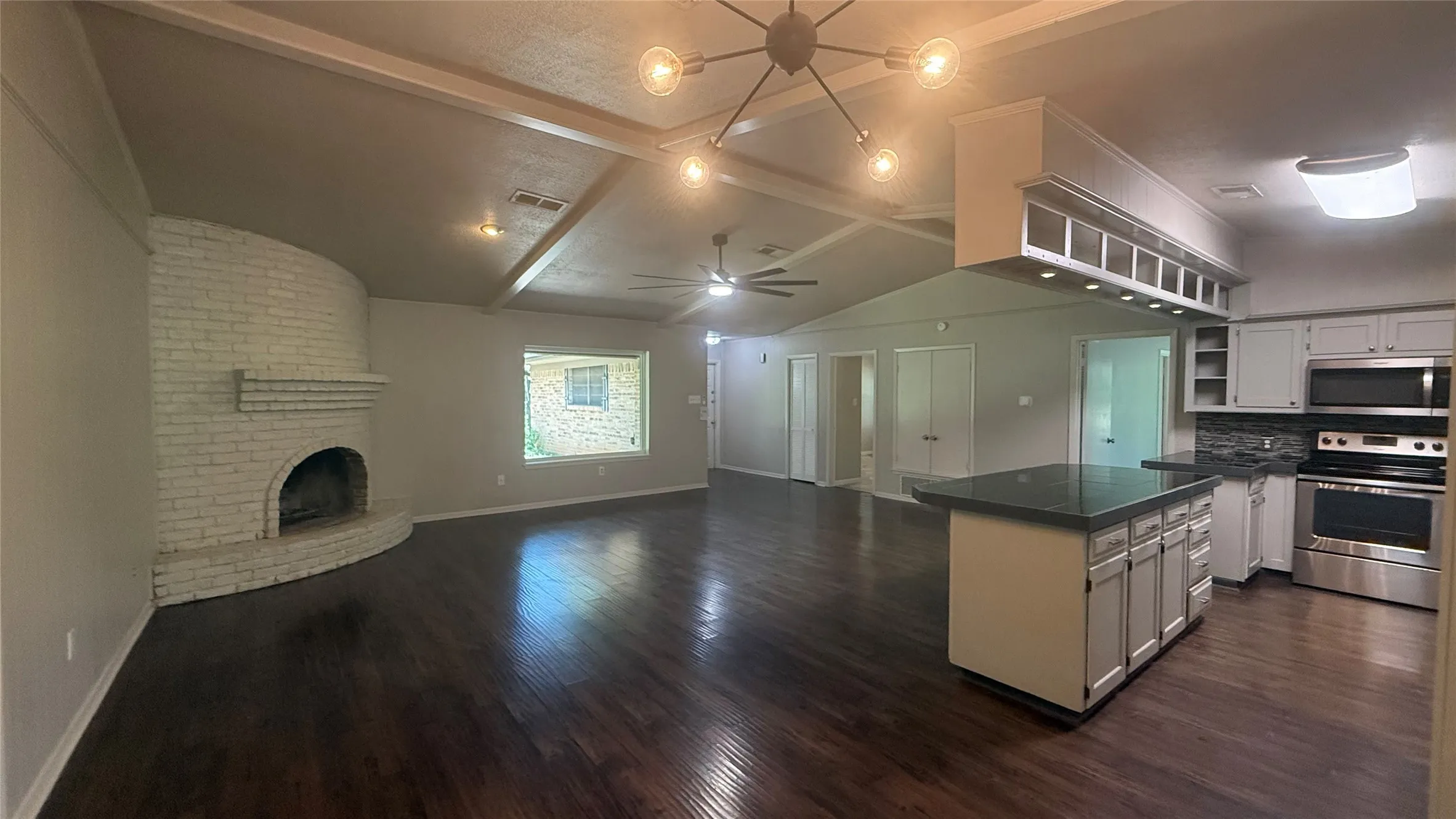 Open-concept interior featuring wood-finish flooring, a white brick fireplace, vaulted ceilings with exposed beams, an island with a dark countertop, and stainless steel kitchen appliances