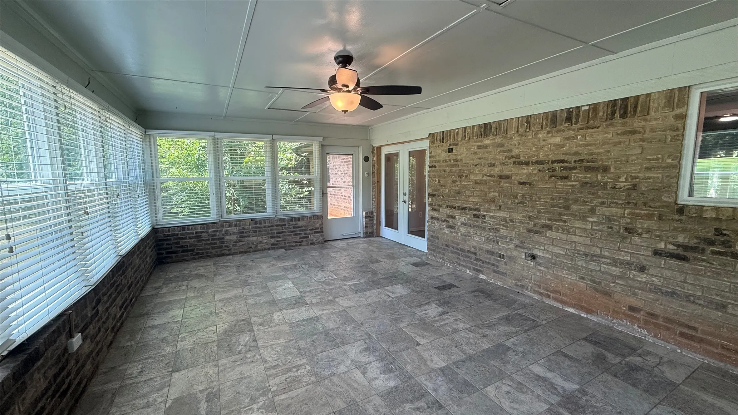 Enclosed patio featuring tile flooring, a ceiling fan with light fixture, multiple windows with blinds, a brick accent wall, and double glass doors
