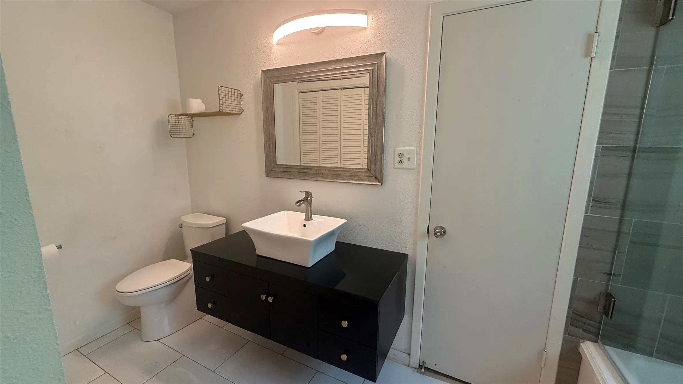 Contemporary bathroom featuring a floating black vanity with a vessel sink, a framed mirror, and light-toned tile flooring