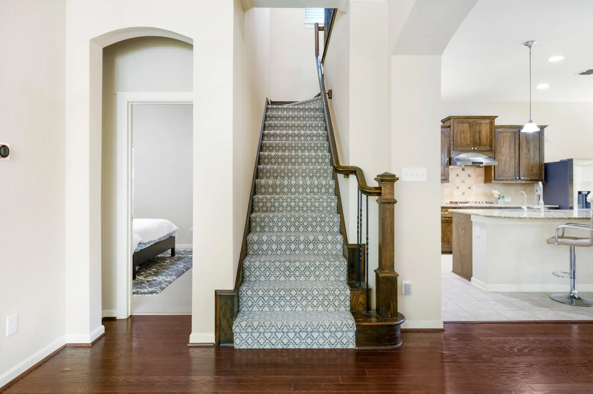 Hardwood flooring extending from the entryway, a staircase with patterned carpeting and wood banister, and a kitchen featuring wood cabinetry, stainless steel appliances, and a breakfast bar with stone countertops
