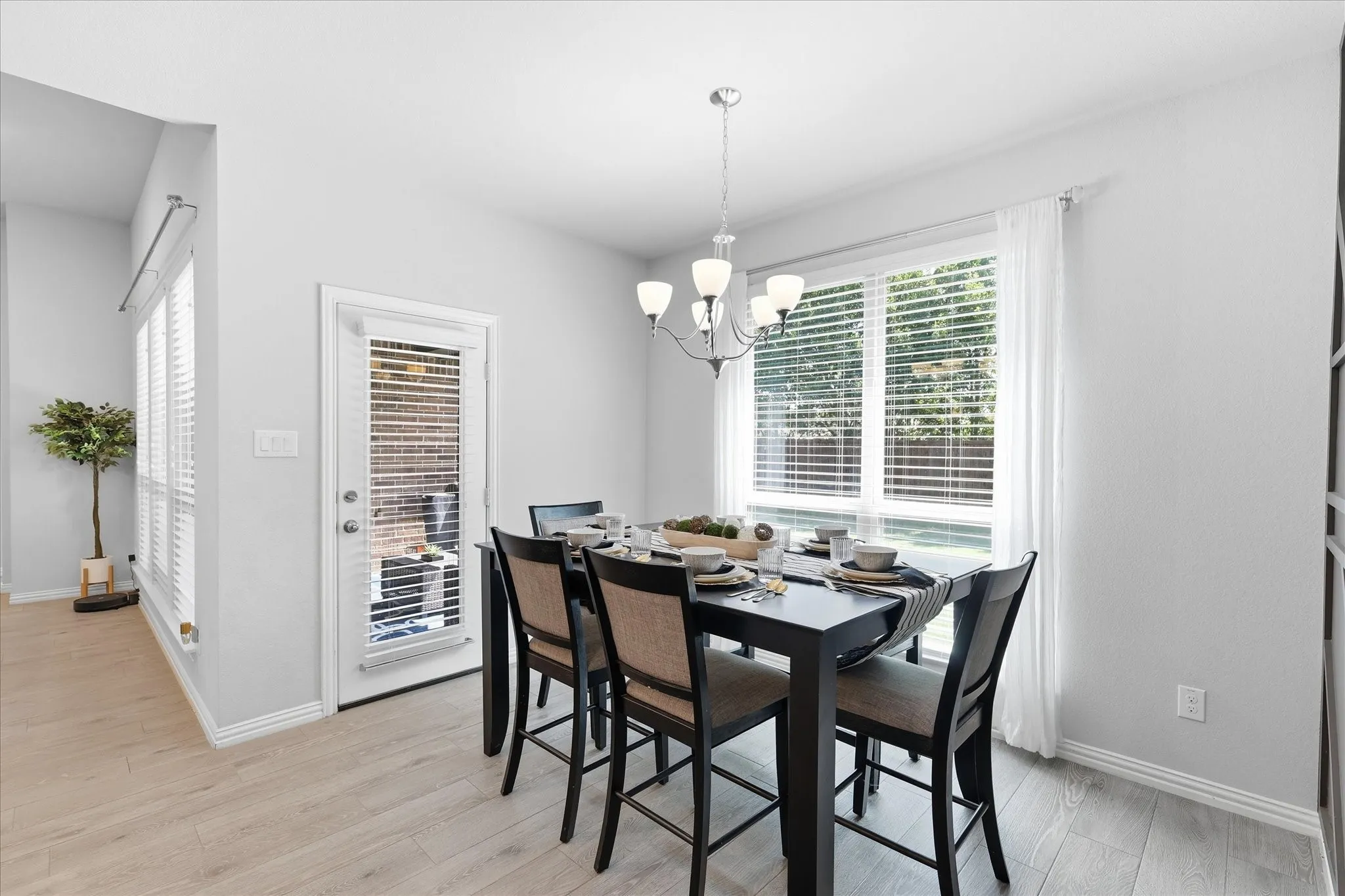 Dining area featuring wood-finish flooring, a contemporary chandelier, large windows with blinds, a white-framed glass door, and crisp white walls