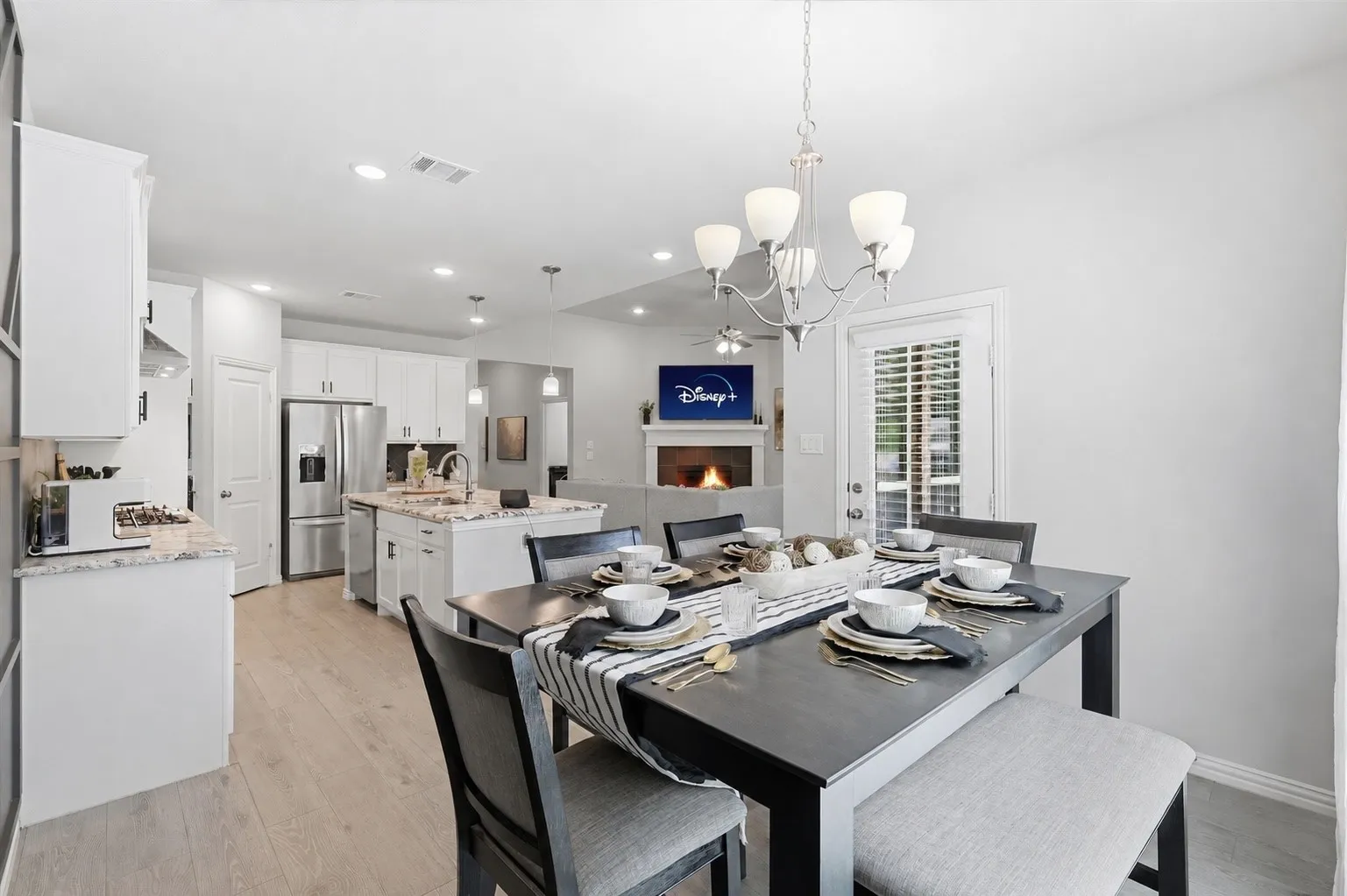 Open-concept living area featuring a formal dining space with chandelier, wood-finish flooring, a kitchen with stainless steel appliances and a center island, and a fireplace with a mounted display