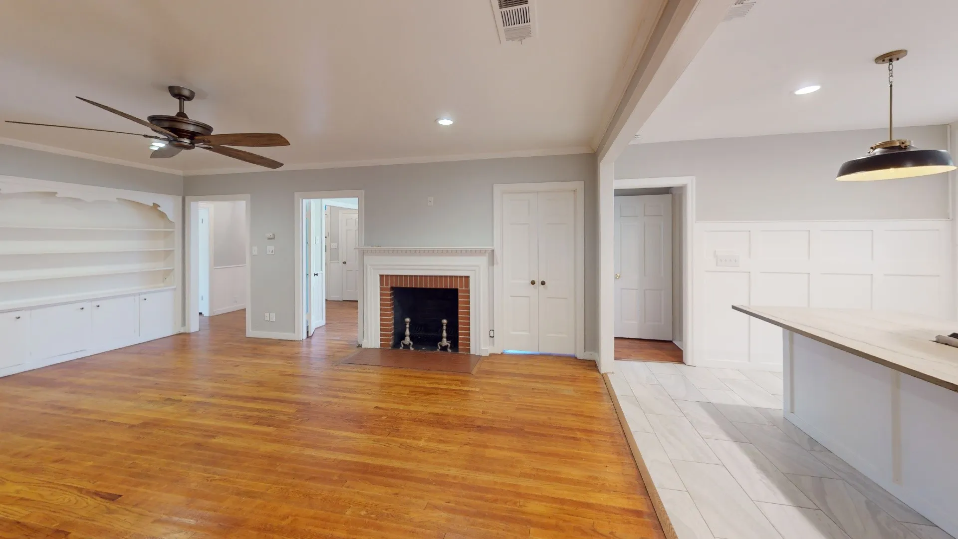 Spacious living area featuring wood-finish flooring, a ceiling fan with integrated lighting, a classic brick fireplace with a white mantel, and built-in shelving with lower cabinetry