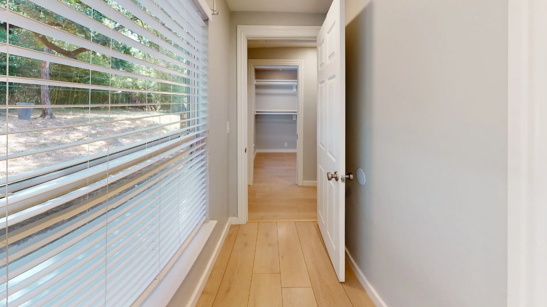 Hallway featuring light wood-finish flooring, neutral wall paint, and a window with horizontal blinds