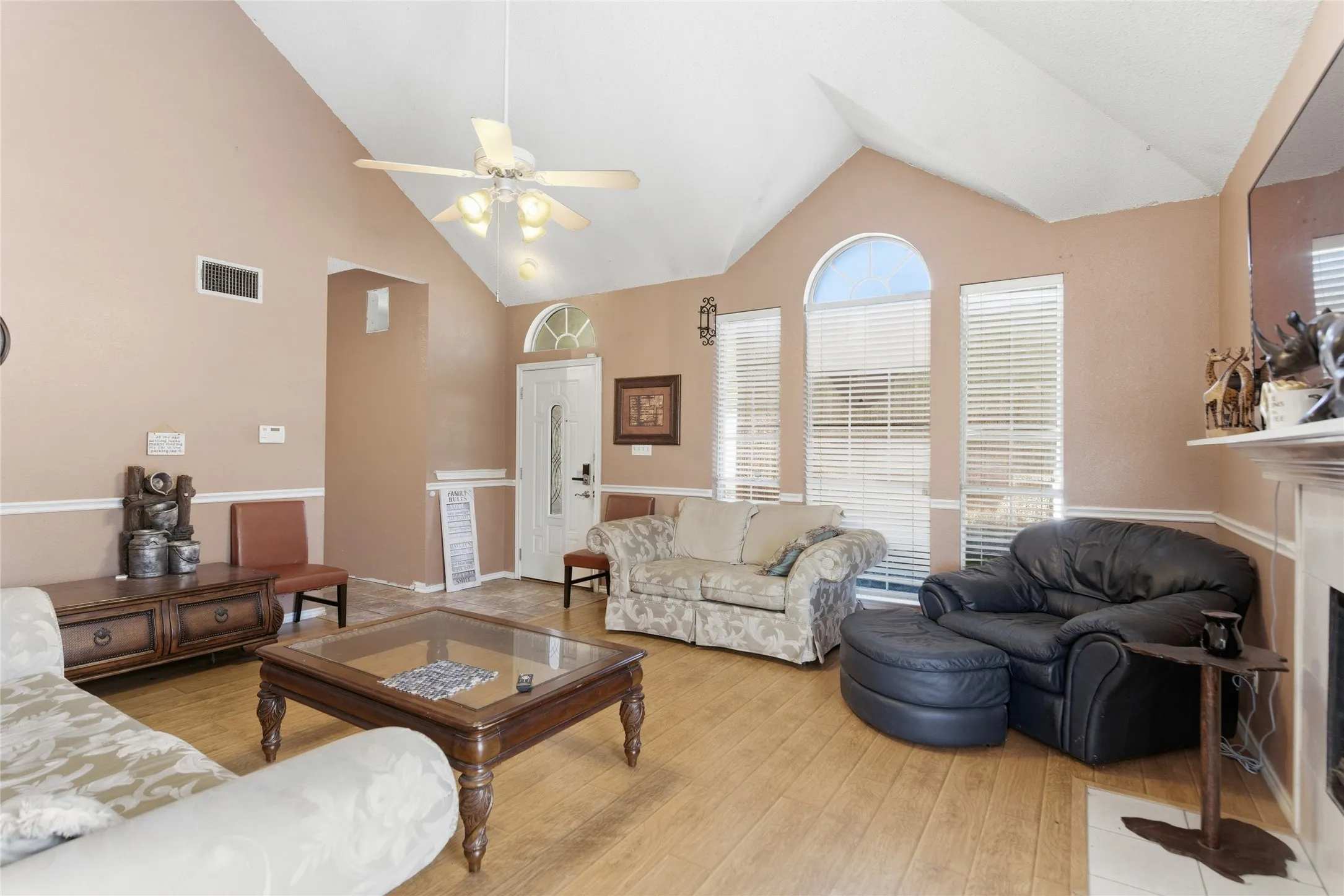 Living area featuring vaulted ceilings, wood-finish flooring, a ceiling fan with integrated lighting, multiple windows with blinds, and a fireplace with a mantel
