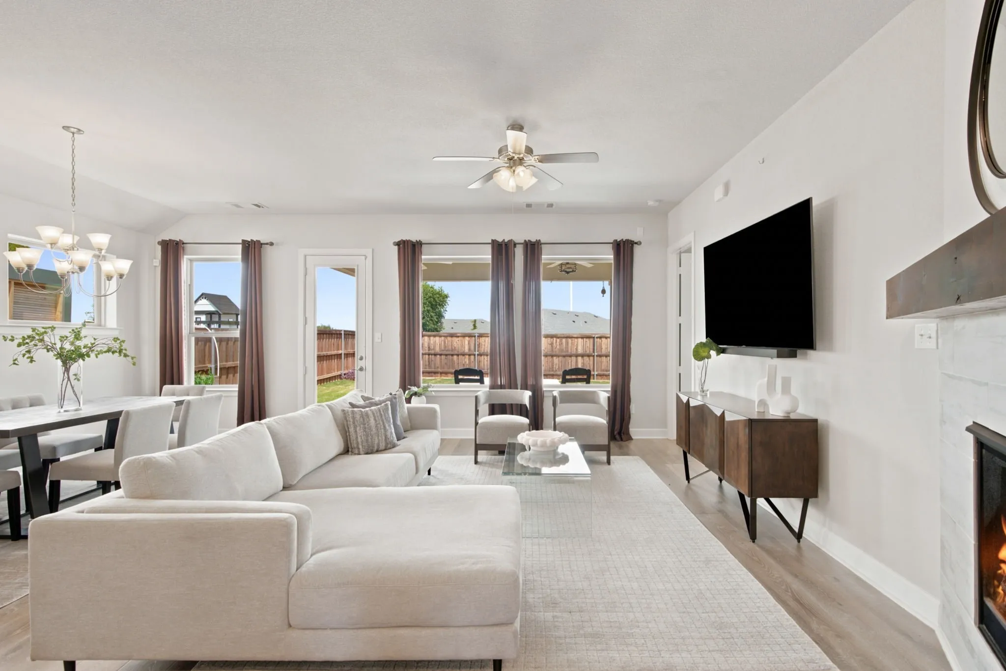 Open-concept living space featuring wood-finish flooring, a white-painted brick fireplace with a wood mantel, a ceiling fan, and multiple large windows for abundant natural light