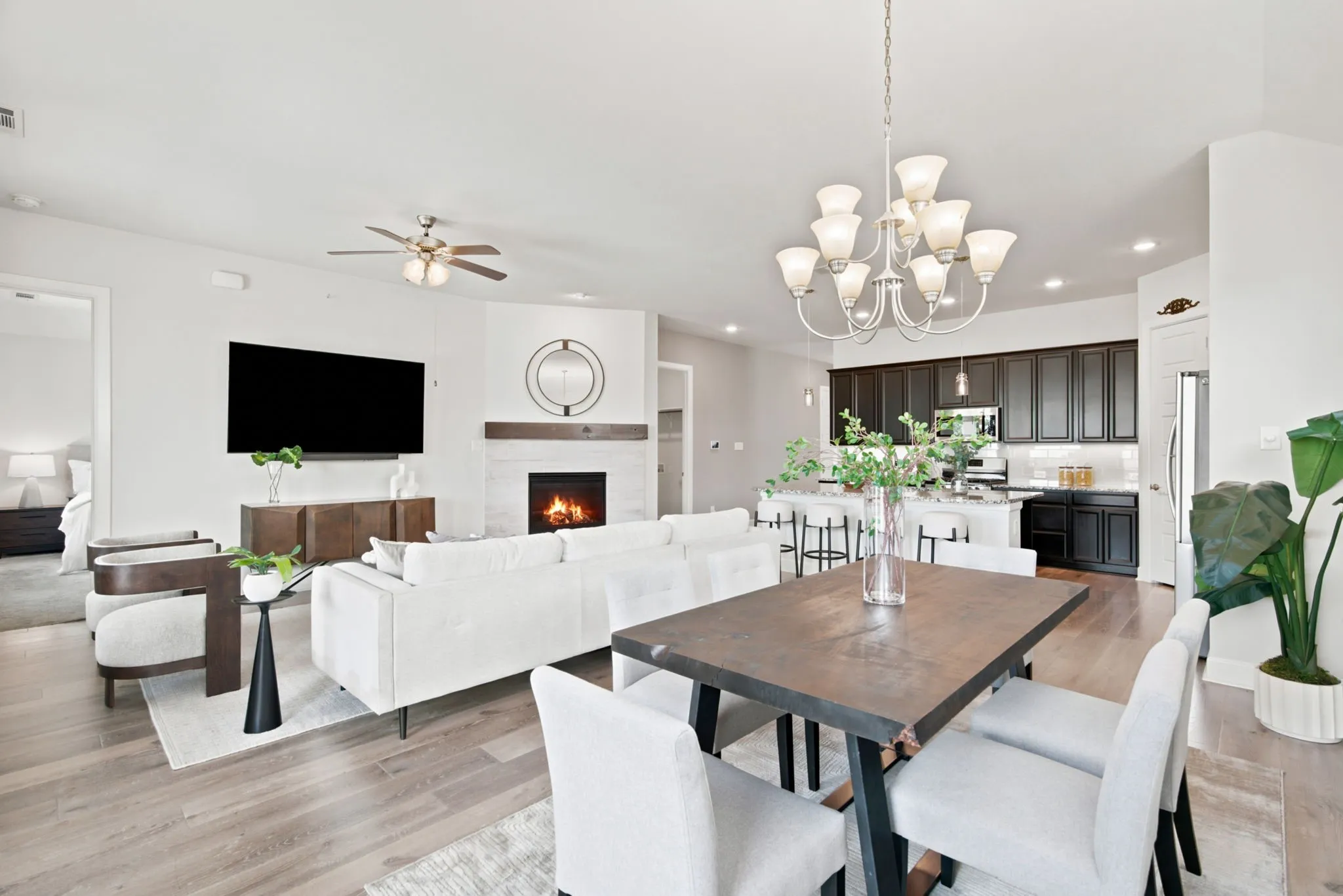 Open-concept living space featuring wood-finish flooring, a dining area with a chandelier, a corner fireplace with a white tile surround and wood mantel, and a kitchen with dark cabinetry and a white island