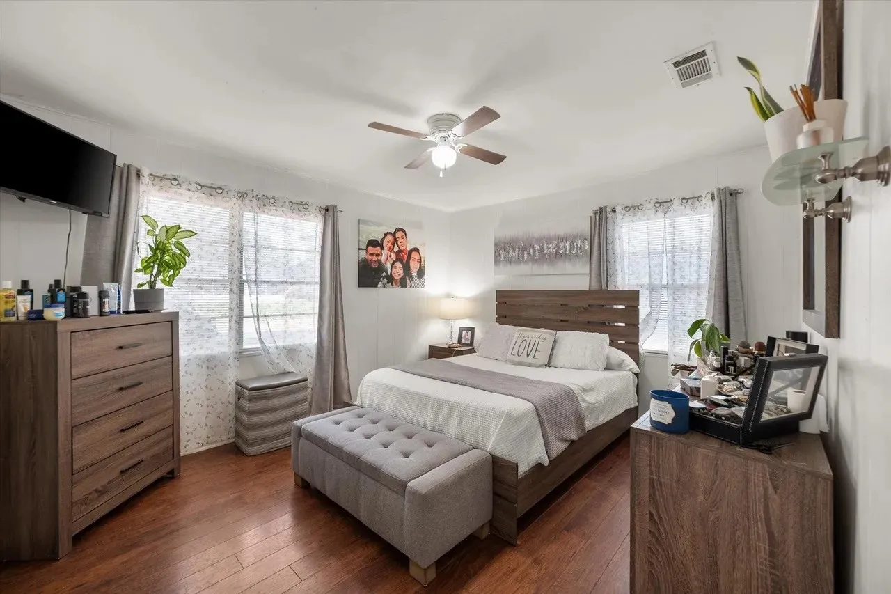 Spacious bedroom featuring wood-finish flooring, two windows with sheer and solid curtains, a ceiling fan with integrated lighting, and a wall-mounted television