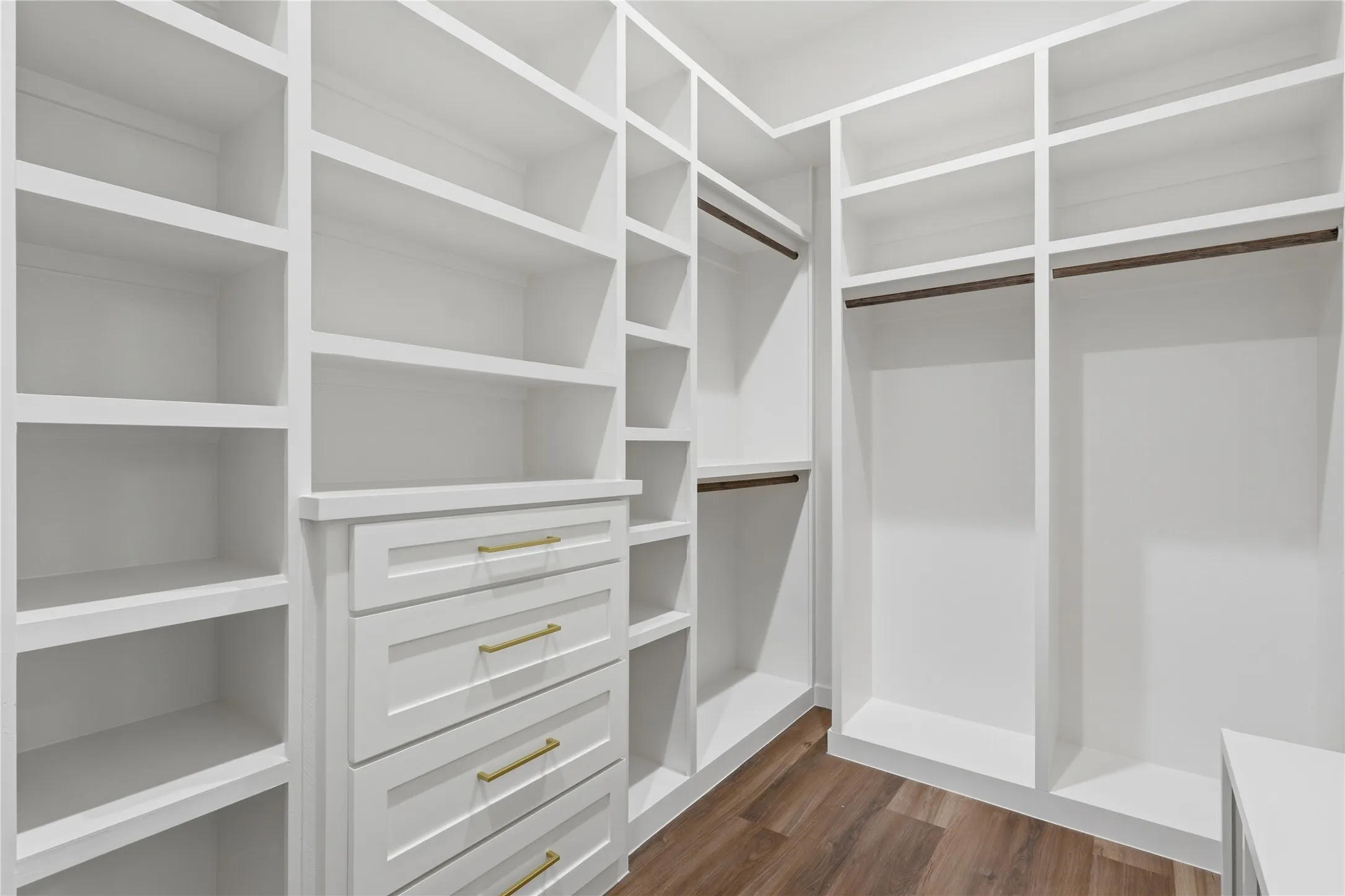 Walk-in closet featuring extensive built-in shelving, multiple hanging rods, three integrated drawers with gold-tone pulls, and wood-finish flooring