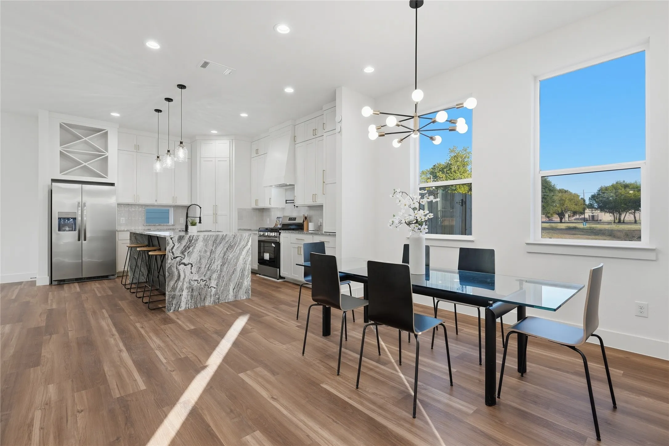 Open-concept living area featuring wood-finish flooring, a kitchen with stainless steel appliances, a large island with a waterfall countertop, white cabinetry, and recessed lighting