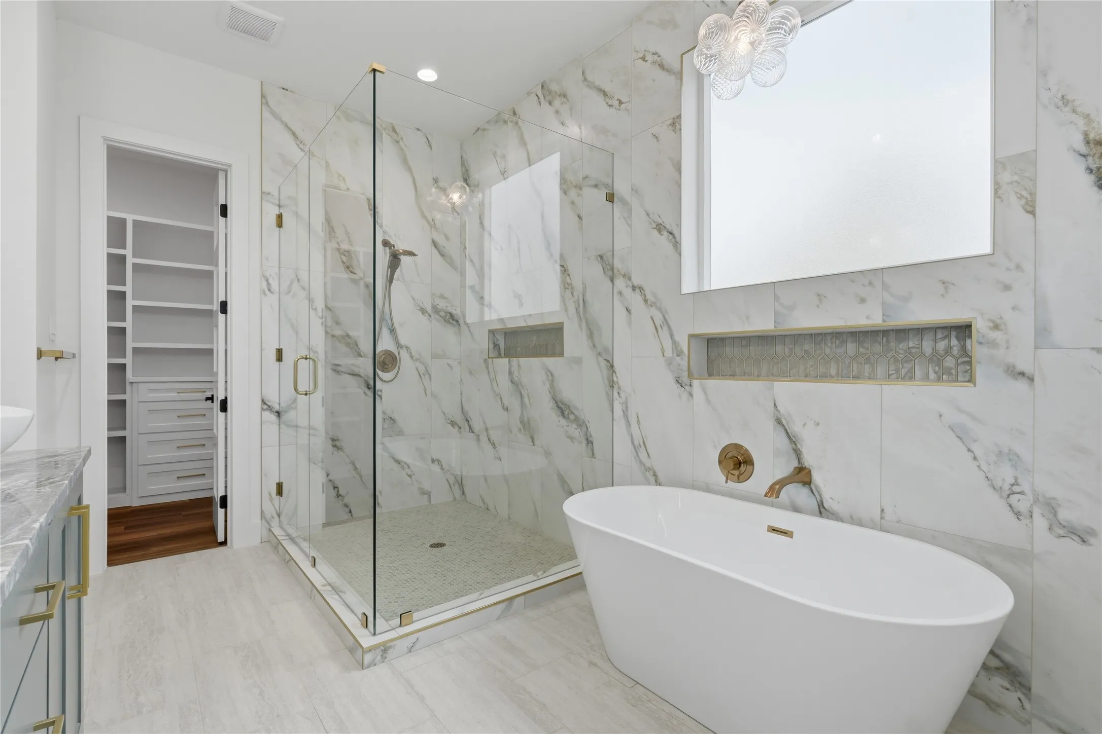 Freestanding soaking tub with brass fixtures, marble-patterned wall tile, and a spacious glass-enclosed shower with mosaic floor