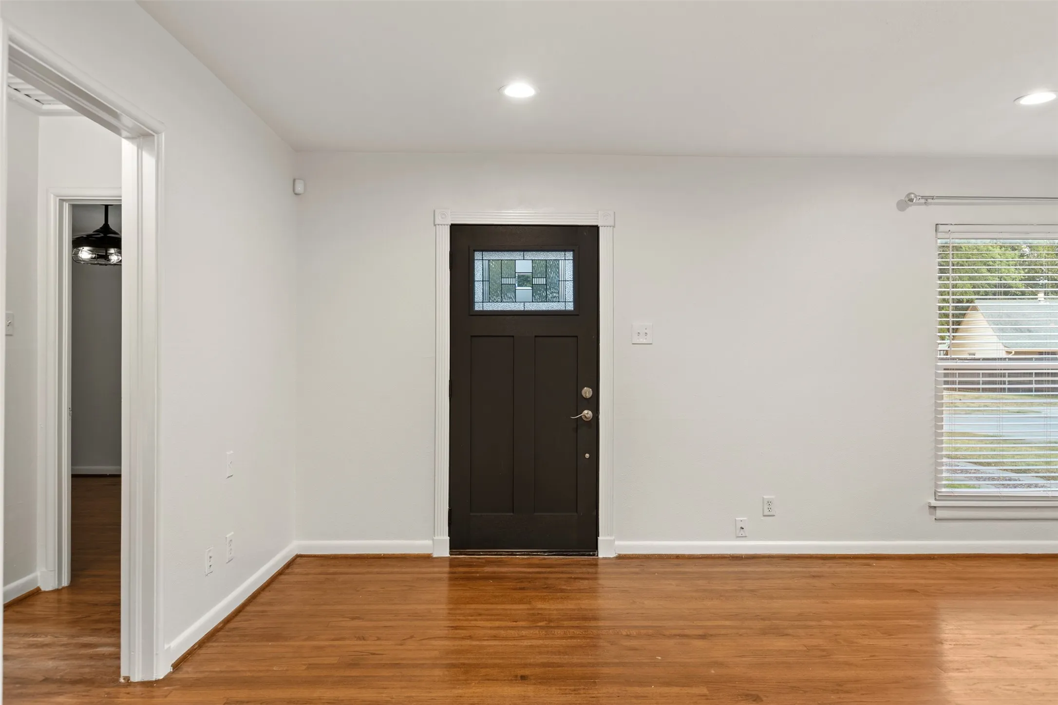 Wood-finish flooring, recessed ceiling lighting, white interior walls, dark paneled entry door with transom window, and a window with blinds