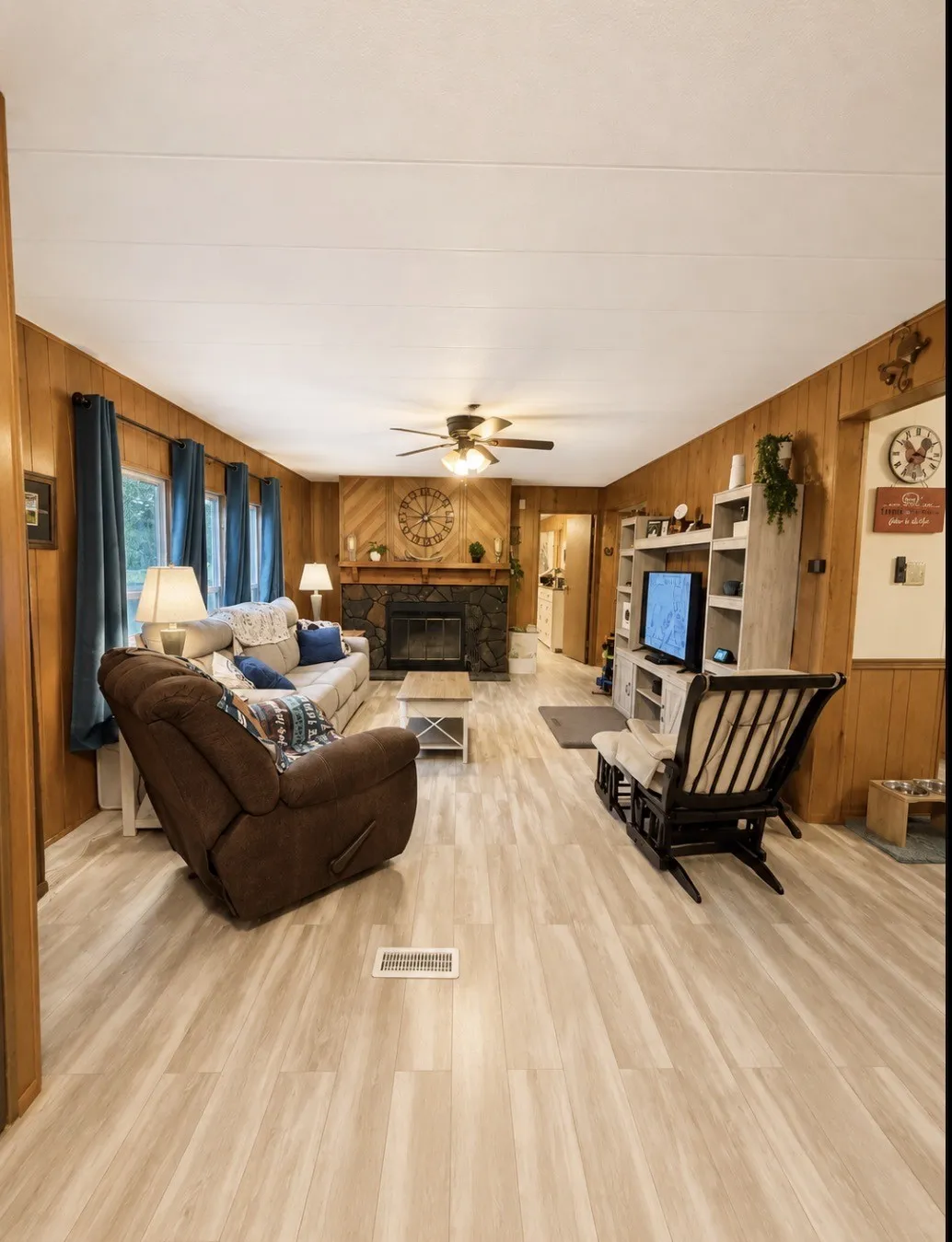 Wood-finish flooring throughout, paneled walls, stone-faced fireplace with wood mantel, and an overhead ceiling fan with light fixture