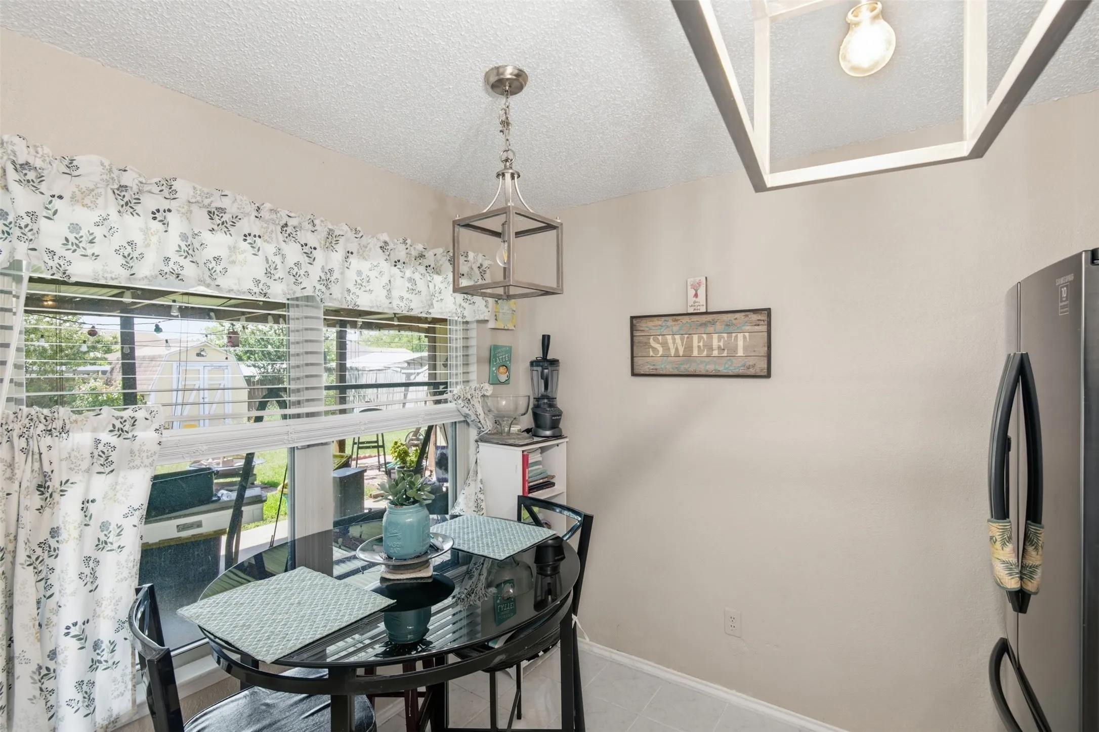 Dining area featuring a contemporary pendant light fixture, stainless steel refrigerator, and a window with horizontal blinds