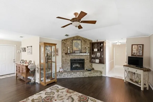 Spacious living area featuring a brick fireplace with a raised hearth, dark wood-finish flooring, a ceiling fan with integrated lighting, and built-in shelving