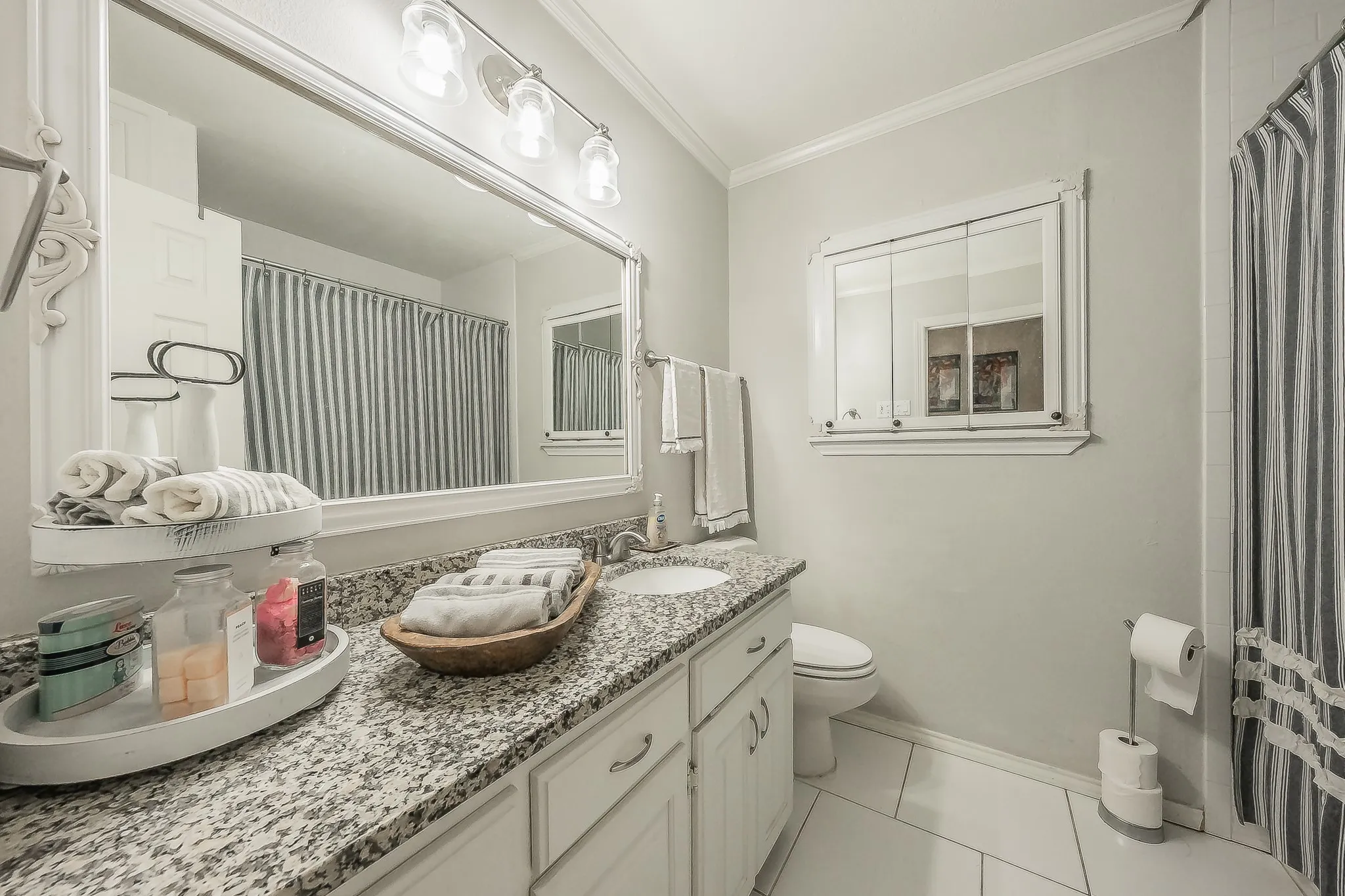 Bathroom featuring a spacious vanity with a granite countertop and dual sinks, white cabinetry, an oversized framed mirror with integrated lighting, and a built-in mirrored medicine cabinet