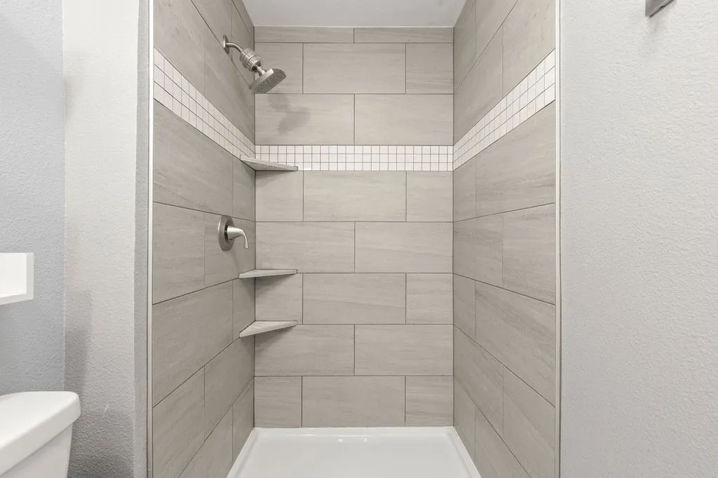 Tiled shower enclosure featuring light grey rectangular tiles, a decorative white mosaic accent band, and a brushed nickel showerhead and control