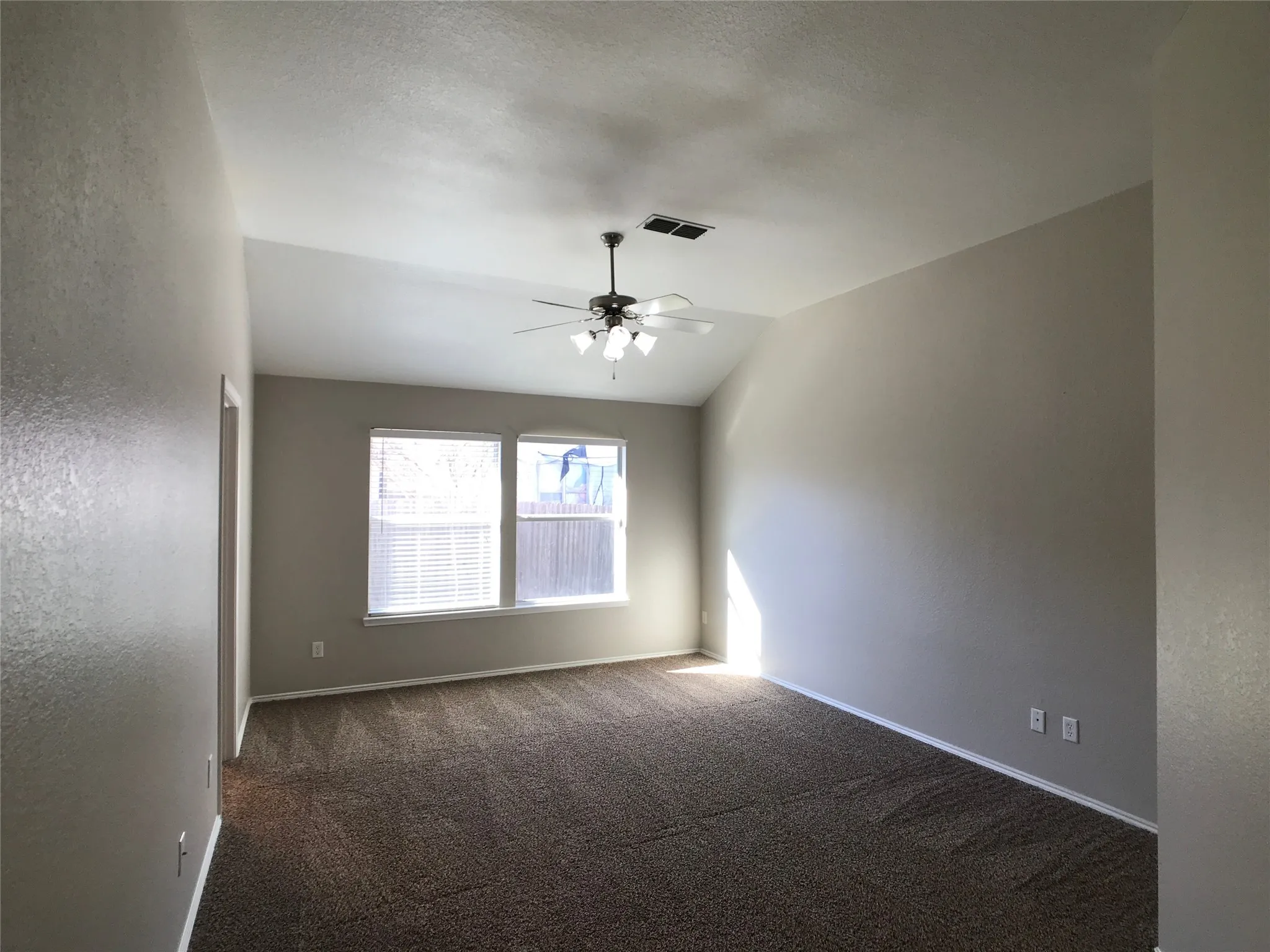 Vaulted ceiling room featuring two windows with horizontal blinds, a ceiling fan with integrated lighting, and patterned brown carpet flooring