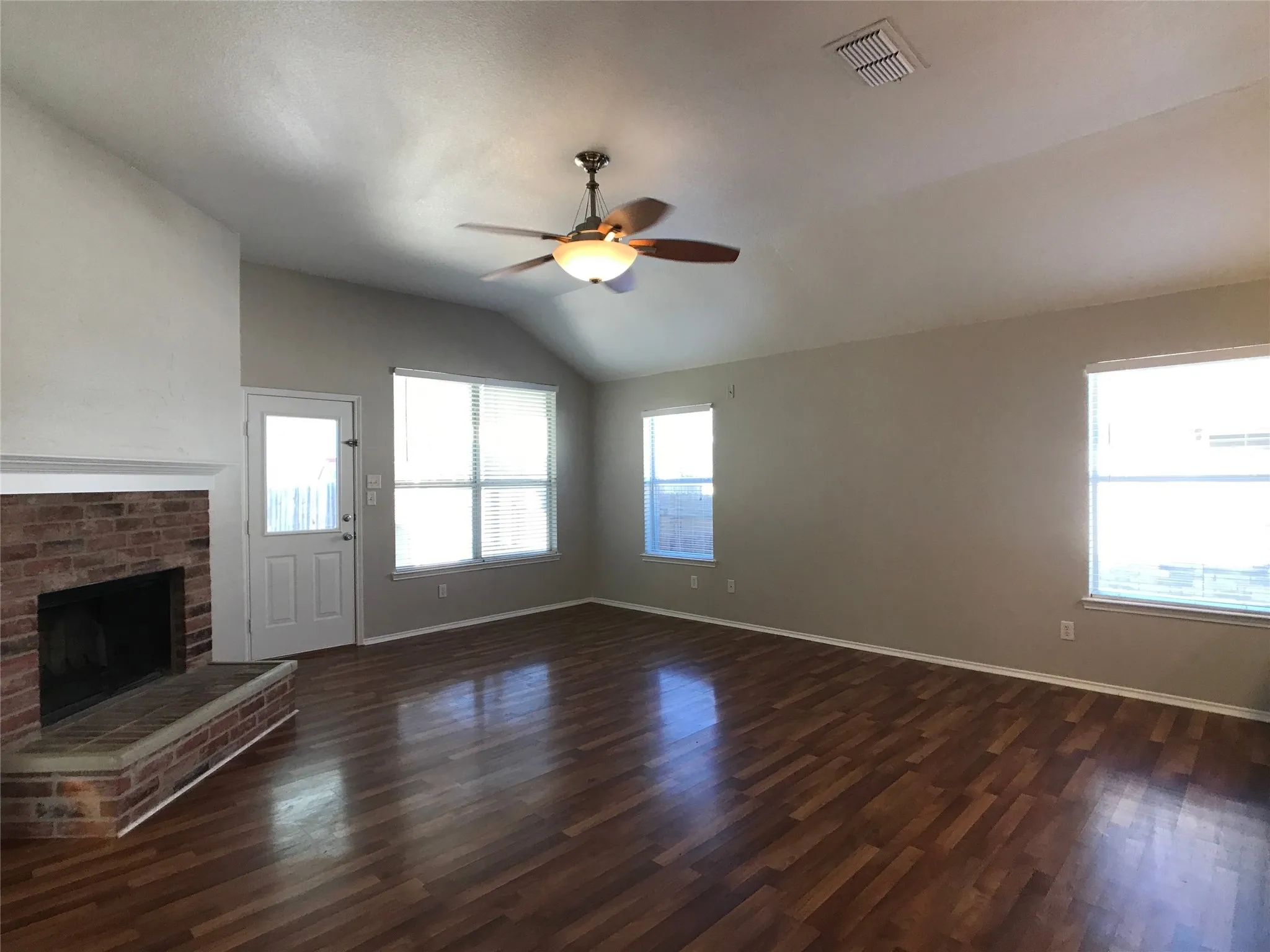 Expansive living area featuring vaulted ceilings, wood-finish flooring, a brick-surround fireplace, multiple windows, and a ceiling fan with integrated lighting