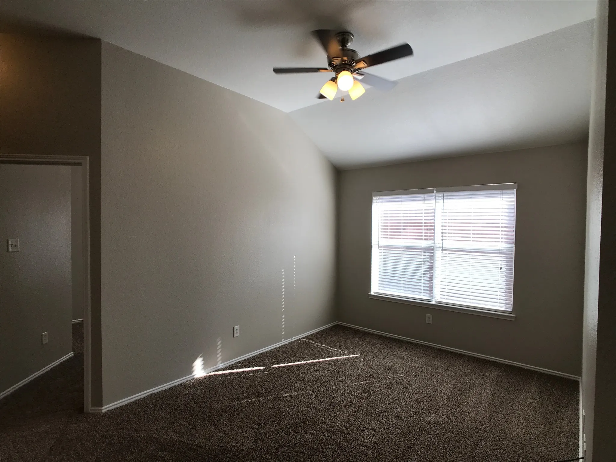Carpeted room featuring a vaulted ceiling with a flush-mounted ceiling fan with integrated lighting, a large window with horizontal blinds, and neutral painted walls