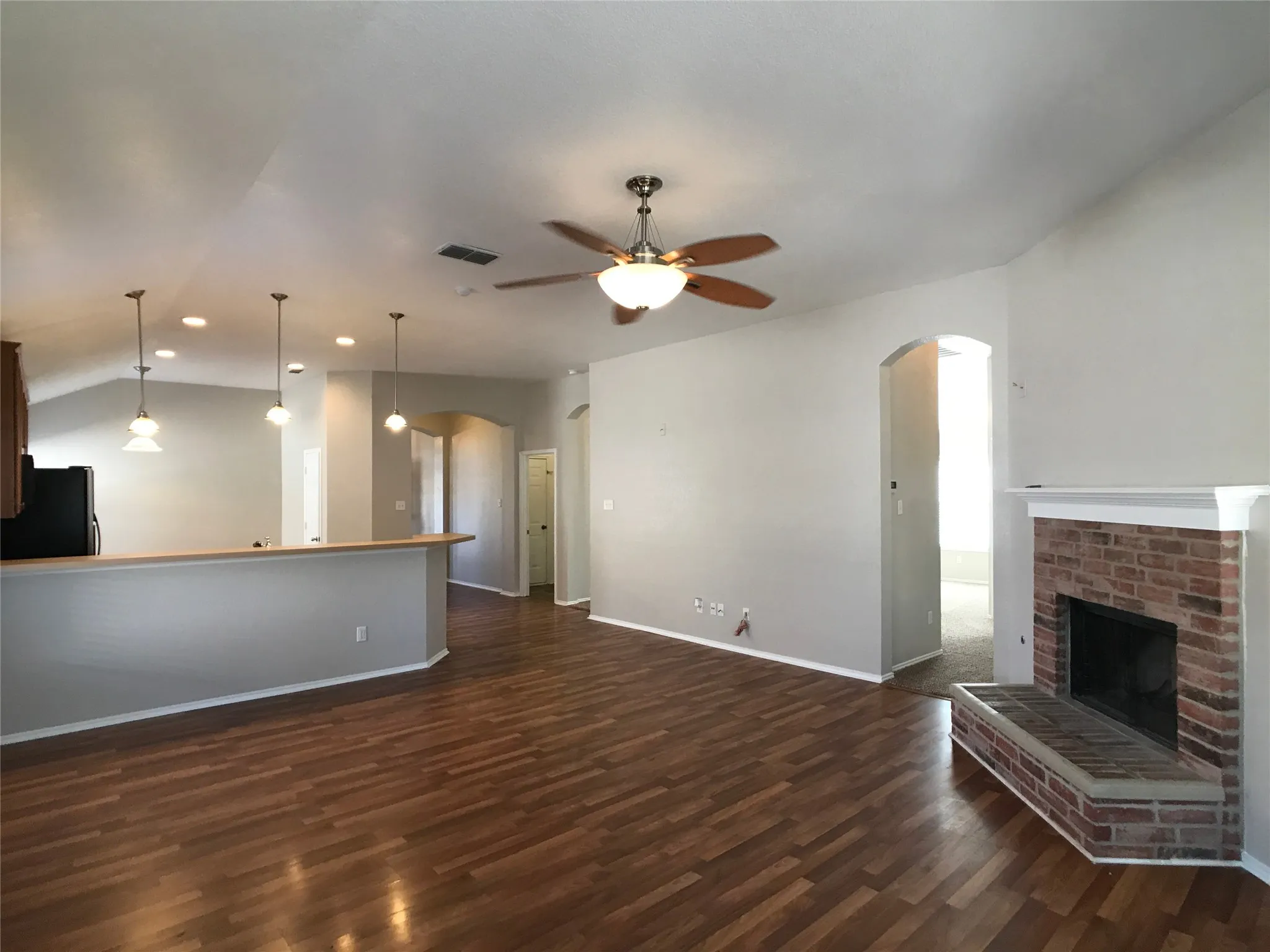 Spacious living area featuring wood-finish flooring, a brick fireplace with a white mantel, and a vaulted ceiling with a ceiling fan