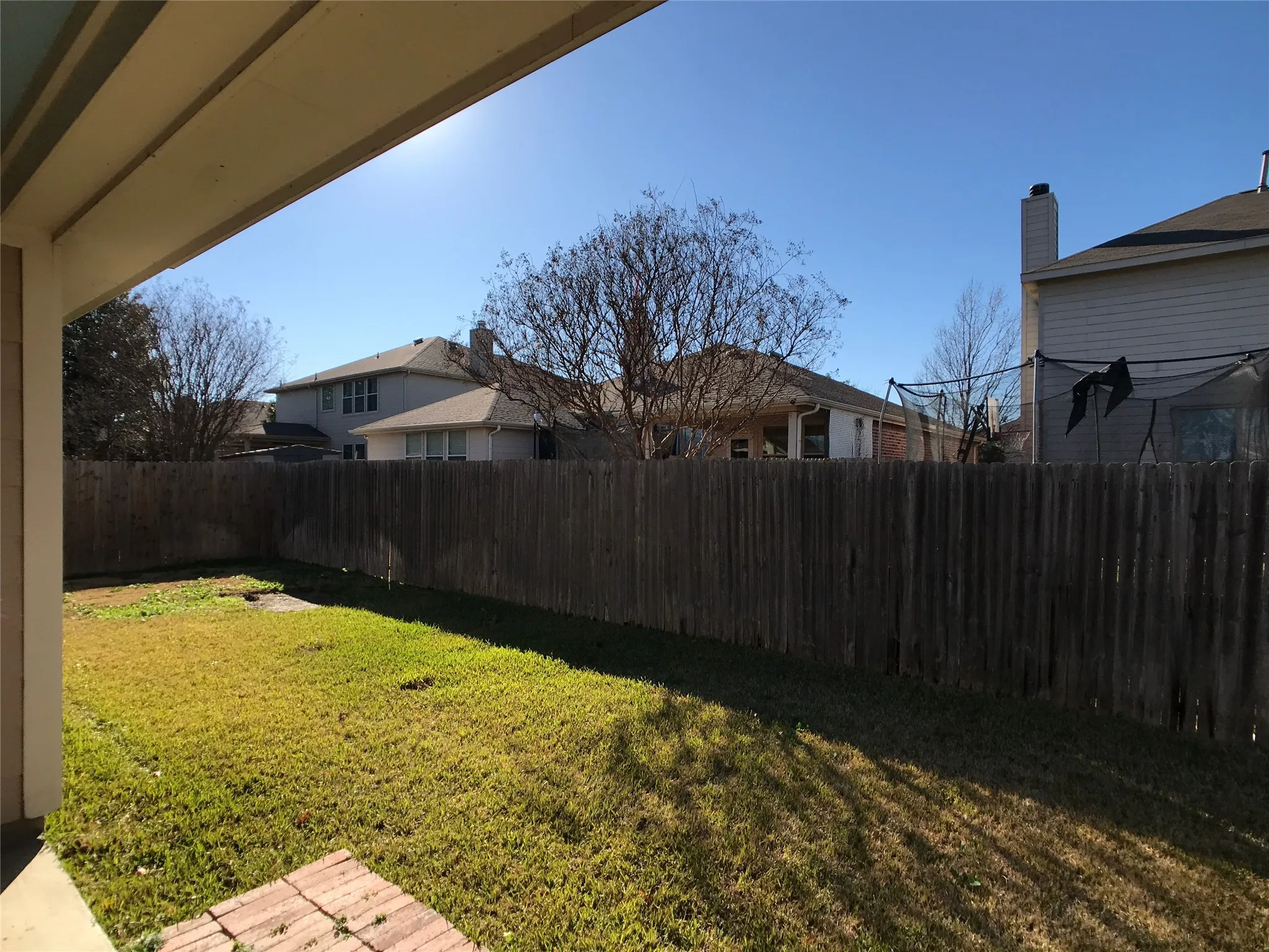 Expansive backyard featuring a wood fence, green lawn, and a covered patio area with a brick paver section