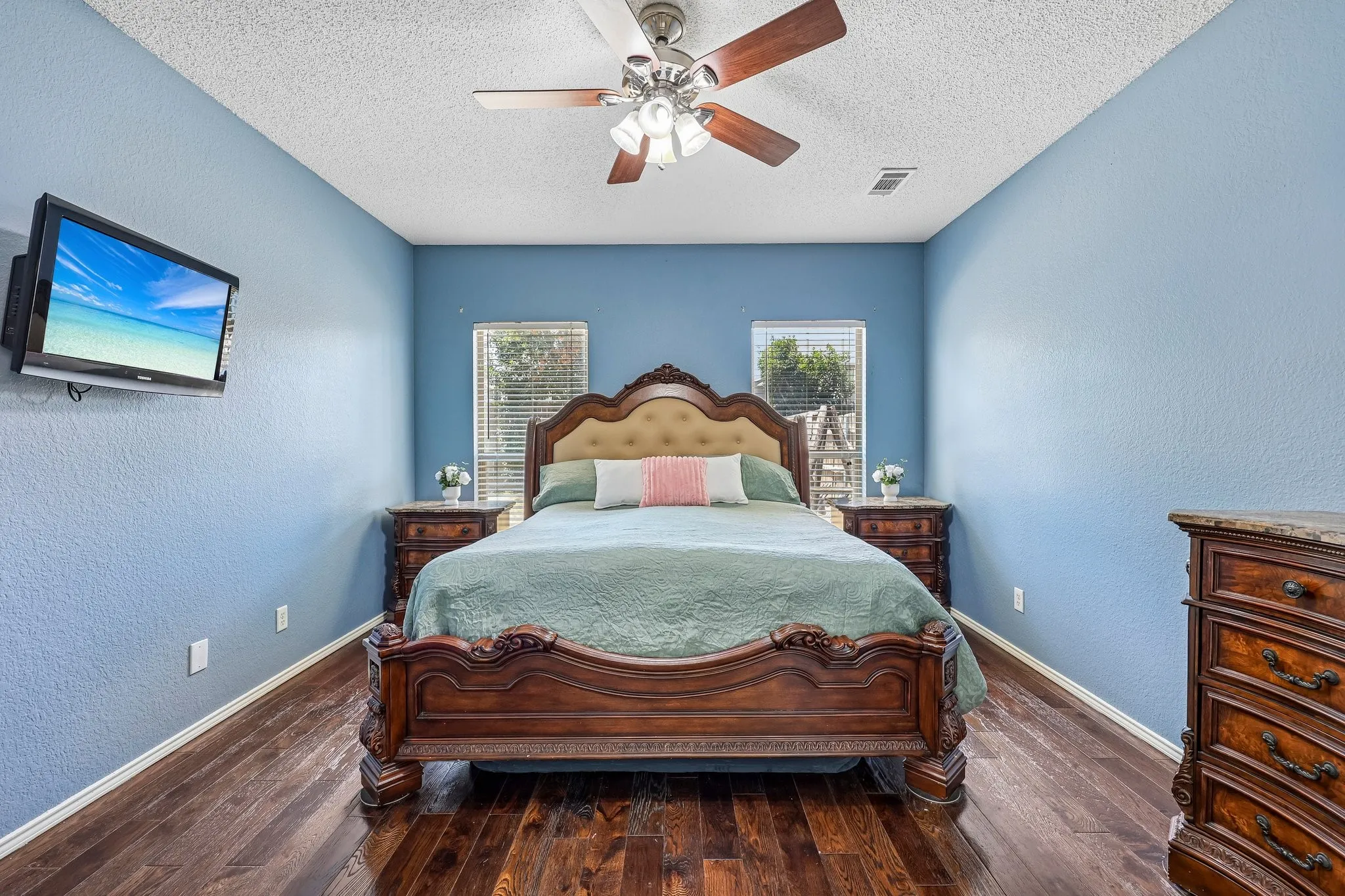 Spacious room featuring rich wood-finish flooring, a ceiling fan with integrated lighting, and two windows