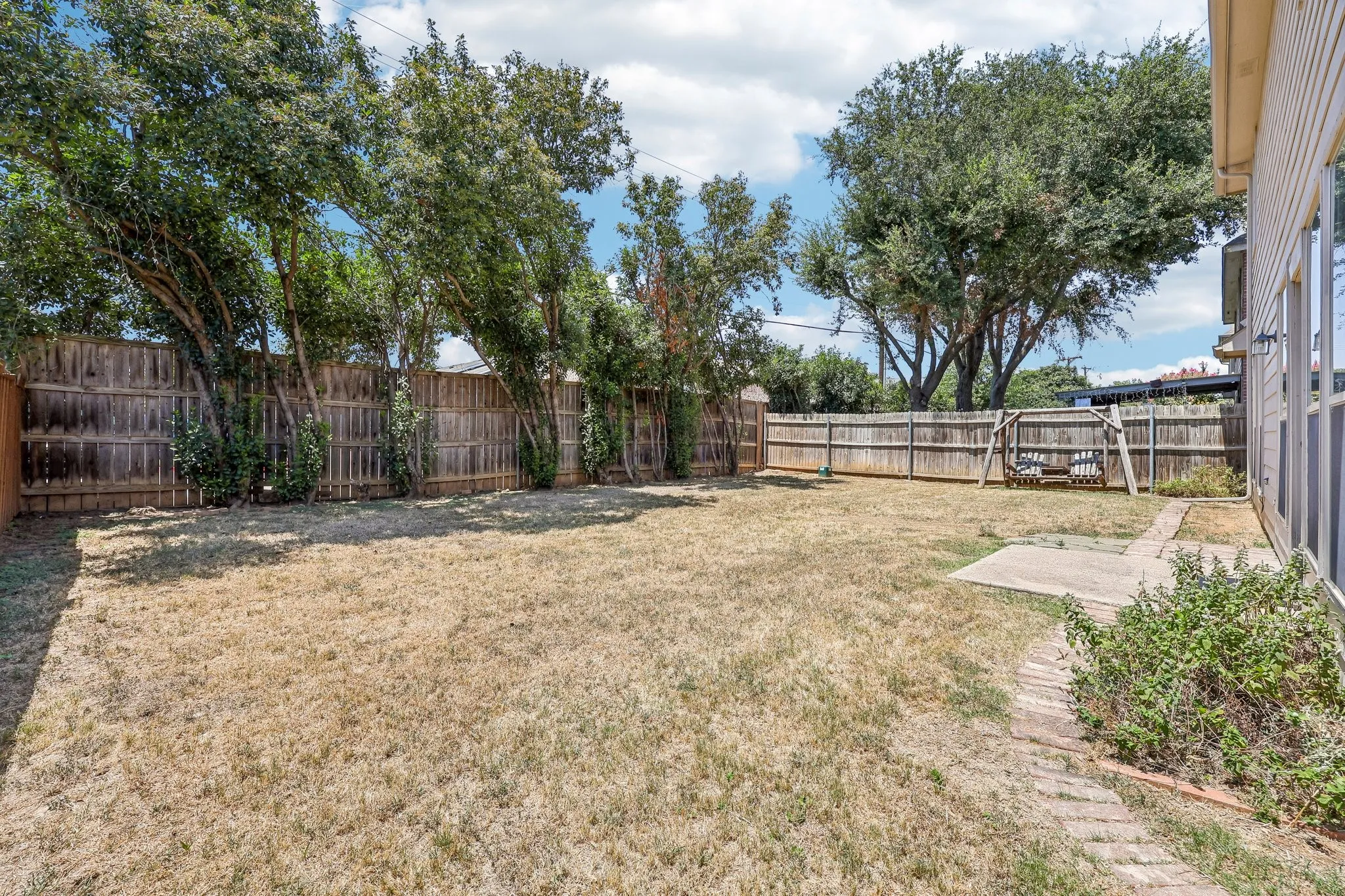 Fenced backyard featuring mature trees, a paved walkway, a concrete patio area, and a swing set