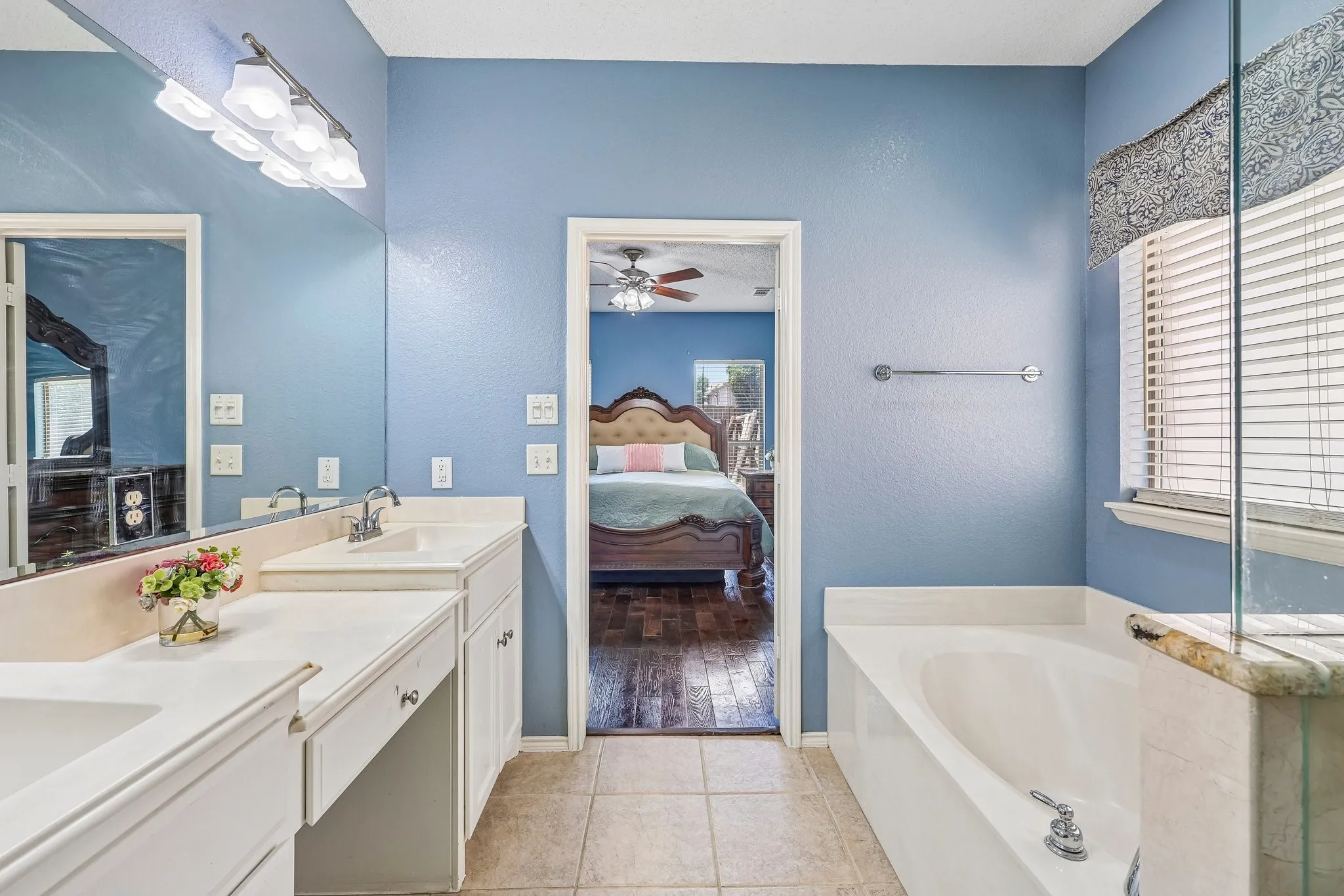 Bathroom featuring a dual vanity with white cabinetry and integrated sinks, a built-in bathtub with tile surround, a window with horizontal blinds, and tile flooring