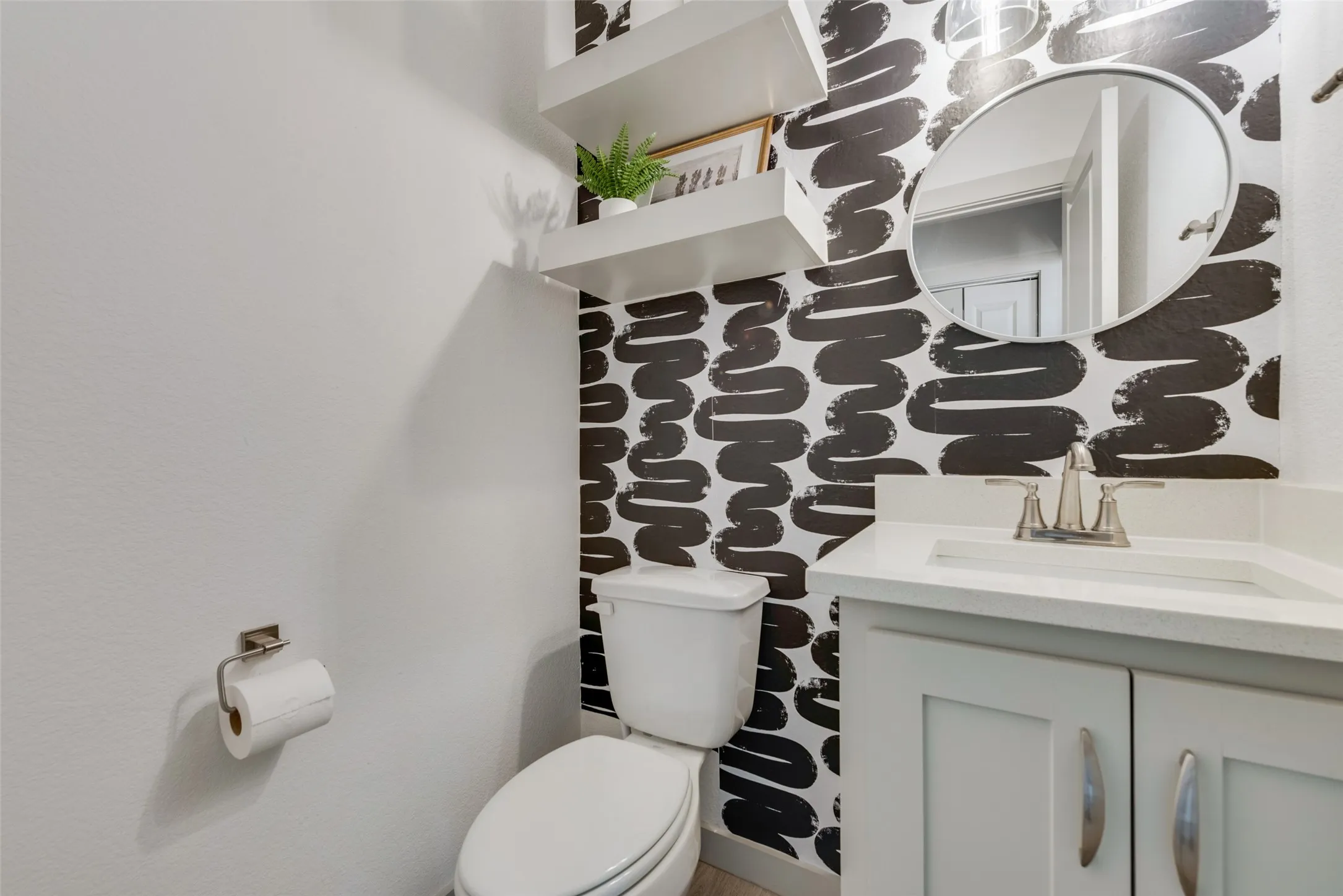 Stylish powder room featuring a striking black and white patterned tile accent wall, round mirror with integrated lighting, white floating vanity with storage cabinetry, white ceramic toilet, and chrome fixtures throughout
