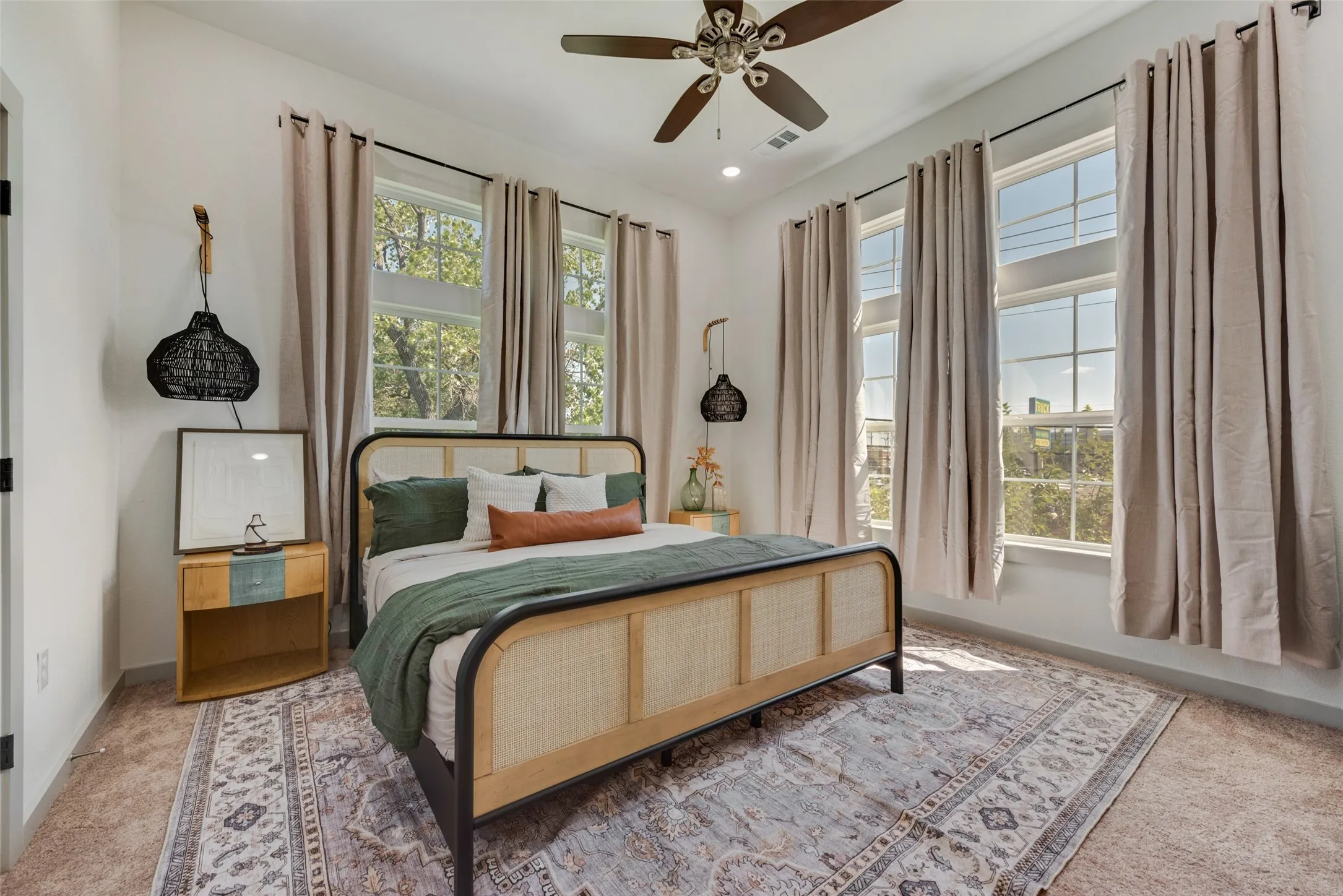 Spacious primary bedroom showcasing a wood-frame bed with rattan paneling, abundant natural light from multiple large windows with floor-to-ceiling drapery, hardwood flooring, an ornate area rug, ceiling fan with integrated lighting, and black pendant fixtures