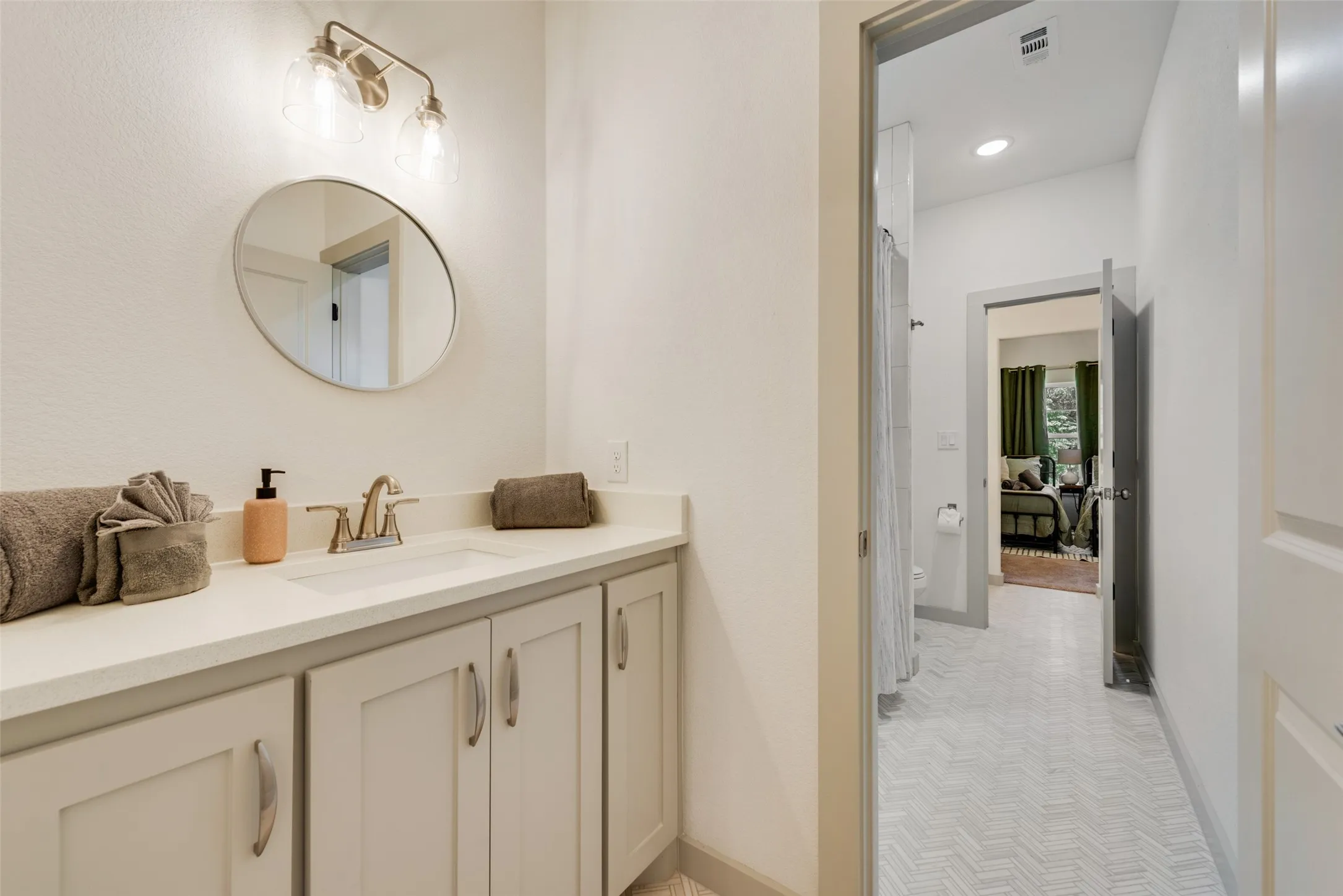 Primary bathroom featuring a cream-finished vanity with dual sinks, brushed gold hardware and faucetry, white quartz countertop, circular framed mirror with brushed gold accent lighting, and access to a spa-like ensuite with tile flooring and glass-enclosed shower