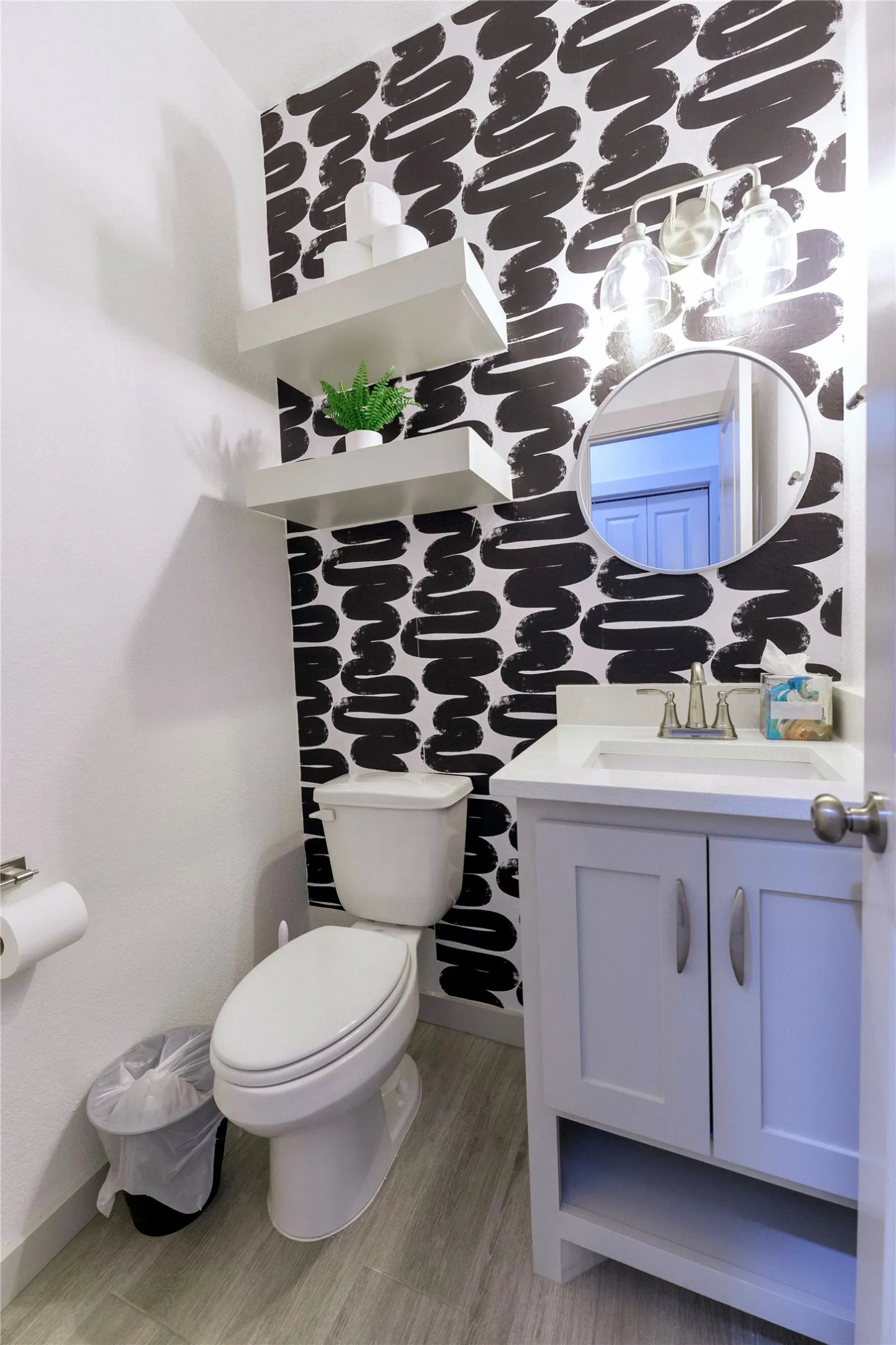 Stylish powder room featuring bold geometric wallpaper in black and white, a round mirror with chrome detailing, and a lavender-painted vanity with cabinet storage