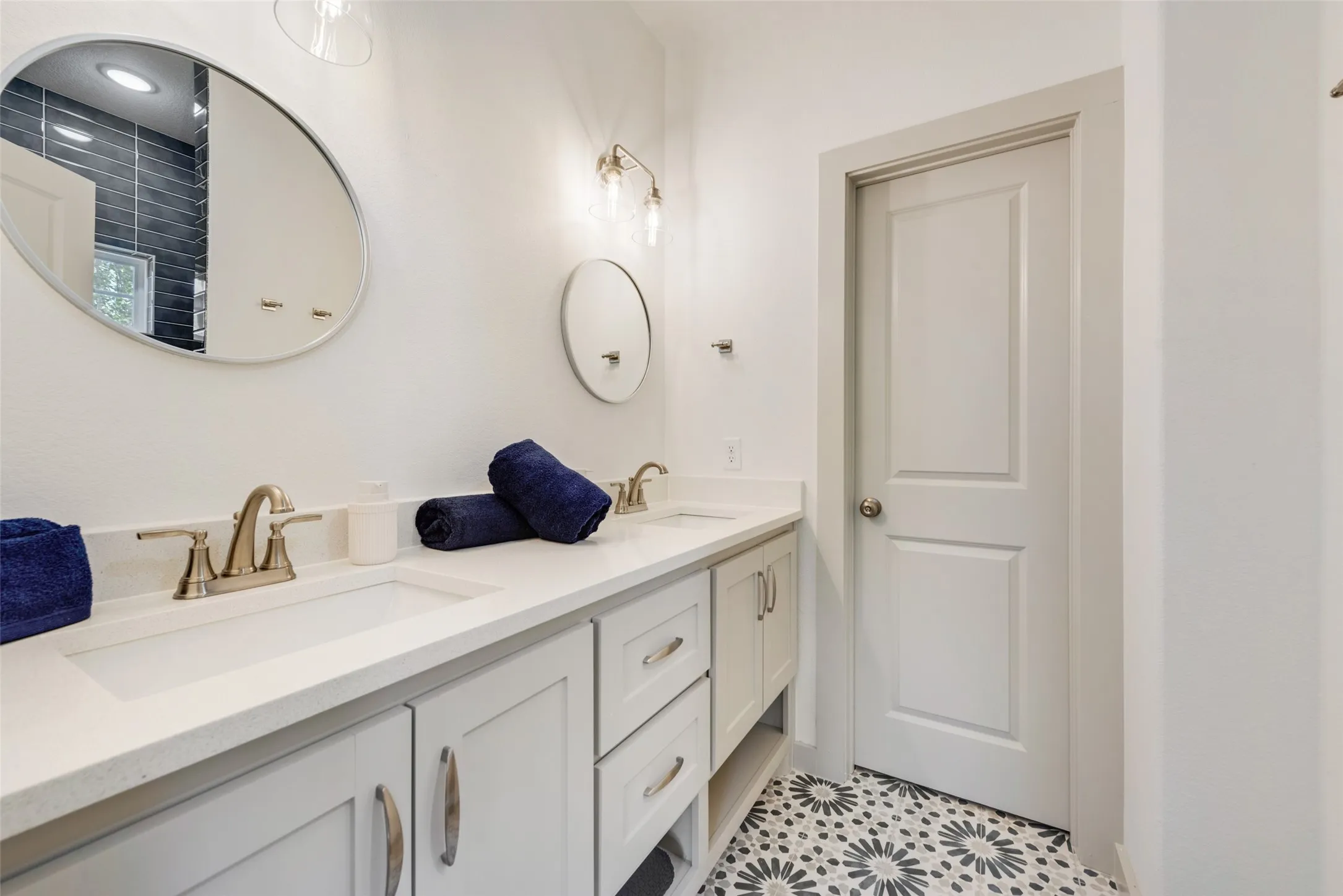 Spa-inspired primary bathroom featuring a lengthy white quartz countertop with dual brass vanity fixtures, soft-close cabinetry with brushed gold hardware, and patterned tile flooring