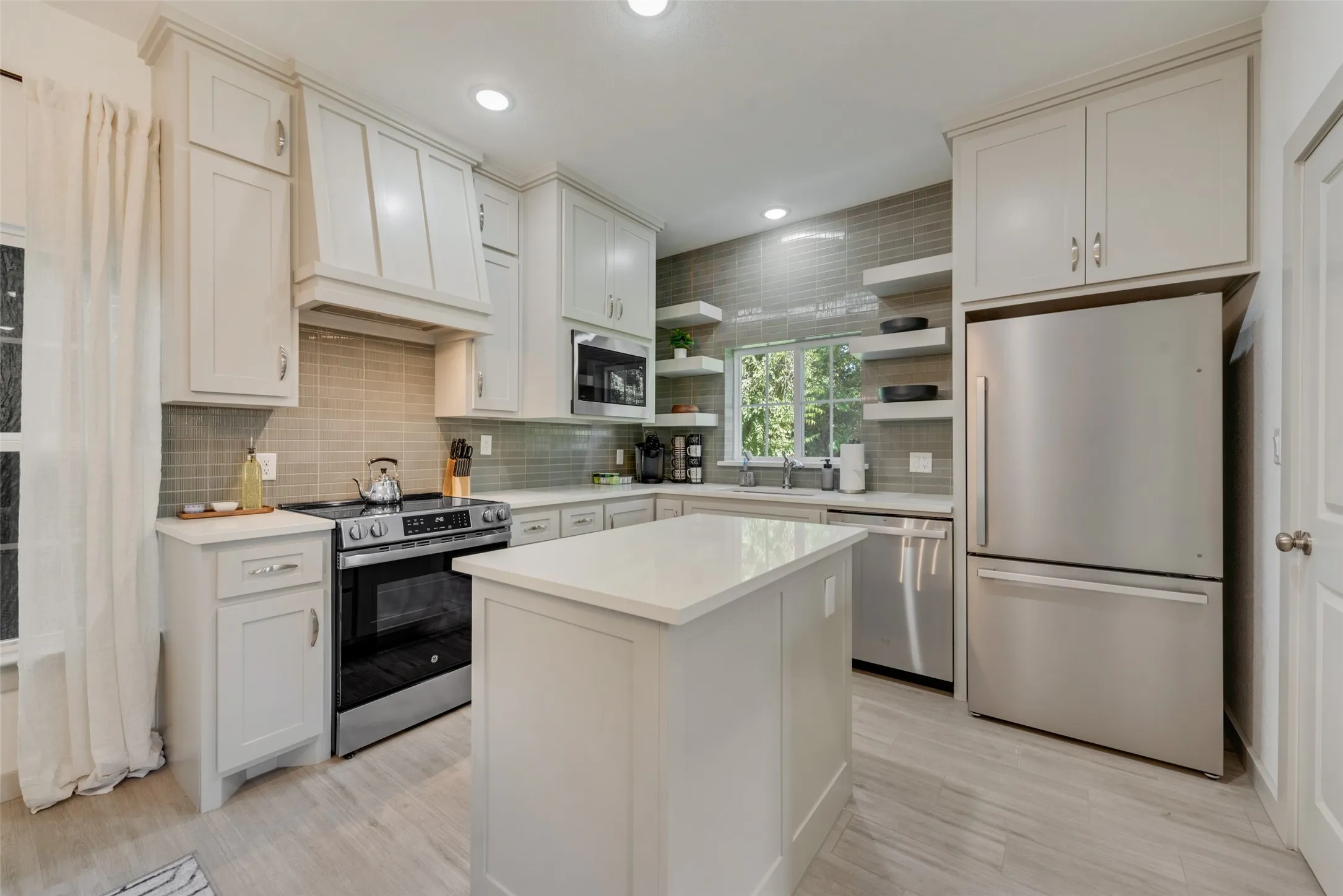 Modern kitchen featuring white cabinetry with shaker-style doors, stainless steel appliances including a black gas range and French-door refrigerator, a large waterfall-edge island with white quartz countertop, gray subway tile backsplash, recessed ceiling lighting, wood-finish flooring, and open shelving with integrated storage throughout the space