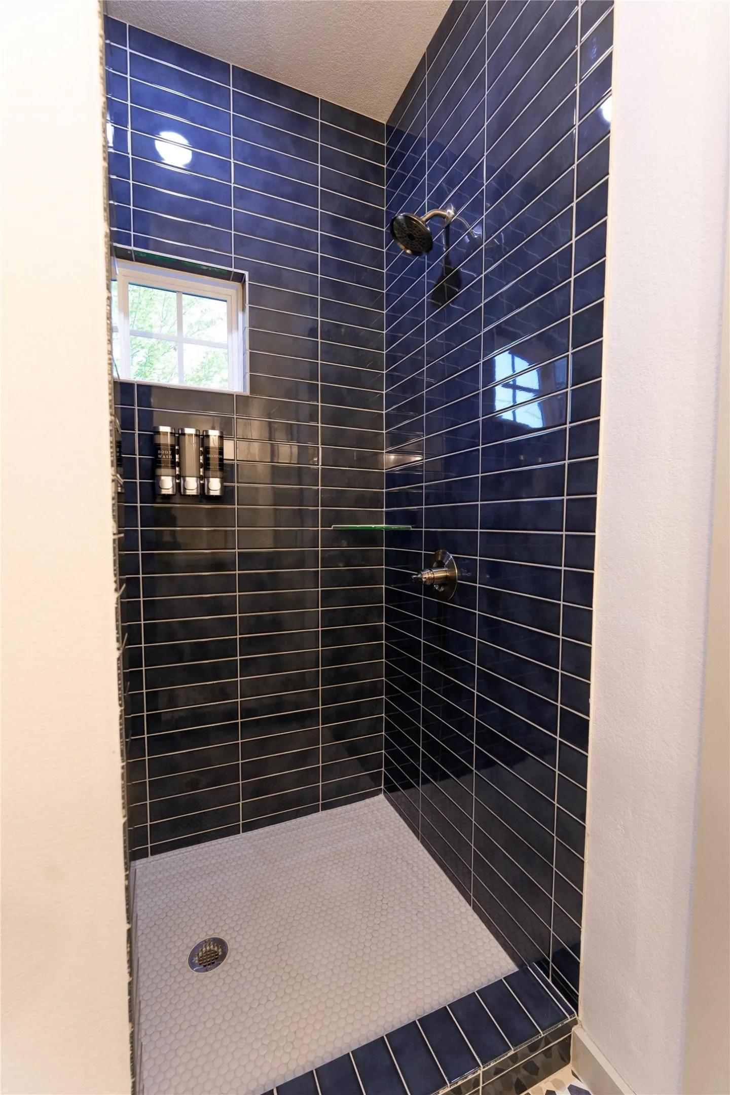 Contemporary walk-in shower featuring horizontal linear tile detailing in deep navy, a frameless glass enclosure, rainfall showerhead, built-in recessed shelving with integrated lighting, and a textured slip-resistant floor base