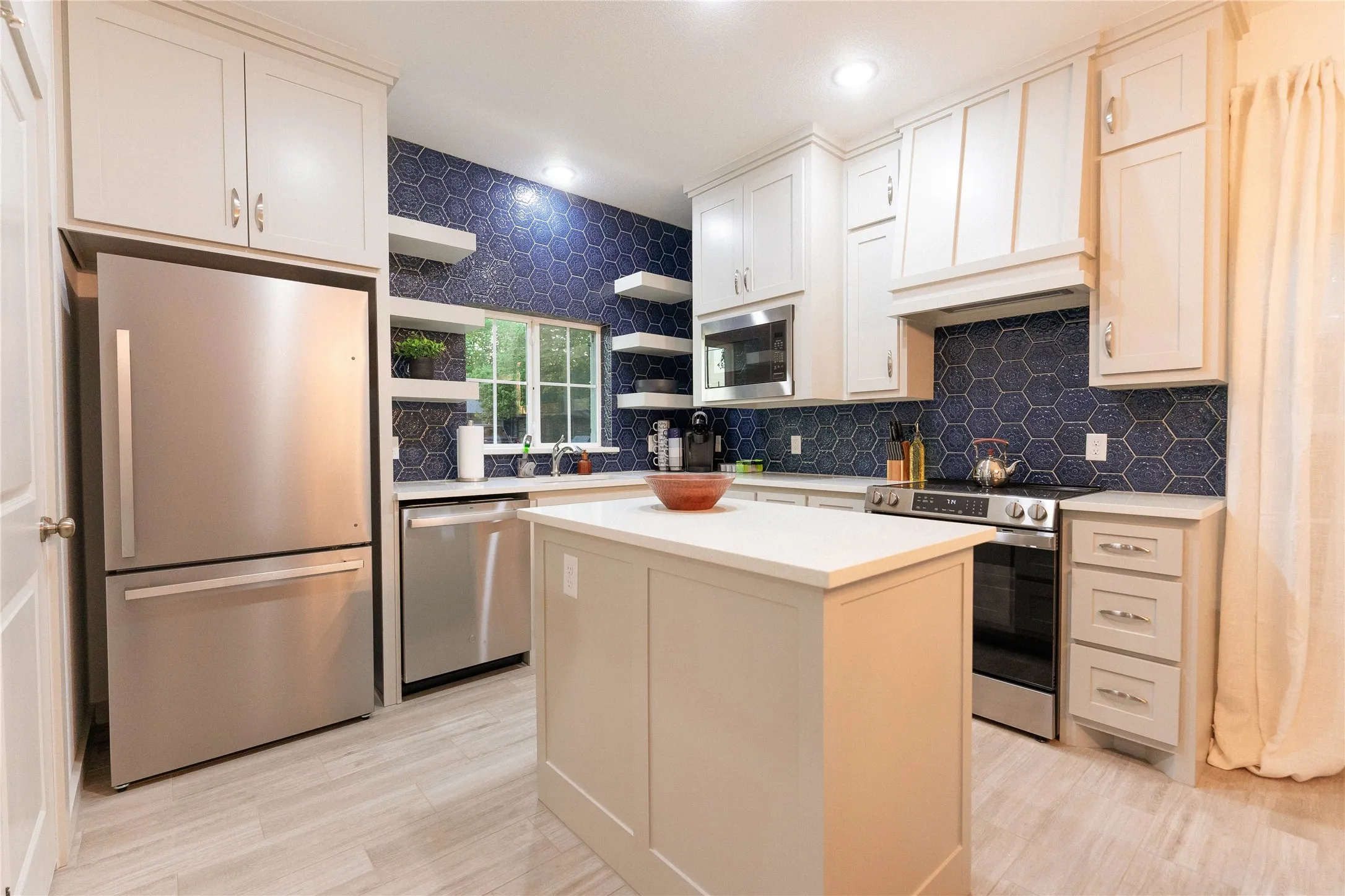 Kitchen featuring a central island, light-toned cabinetry, stainless steel appliances, a deep blue hexagonal tile backsplash, and light wood-finish flooring