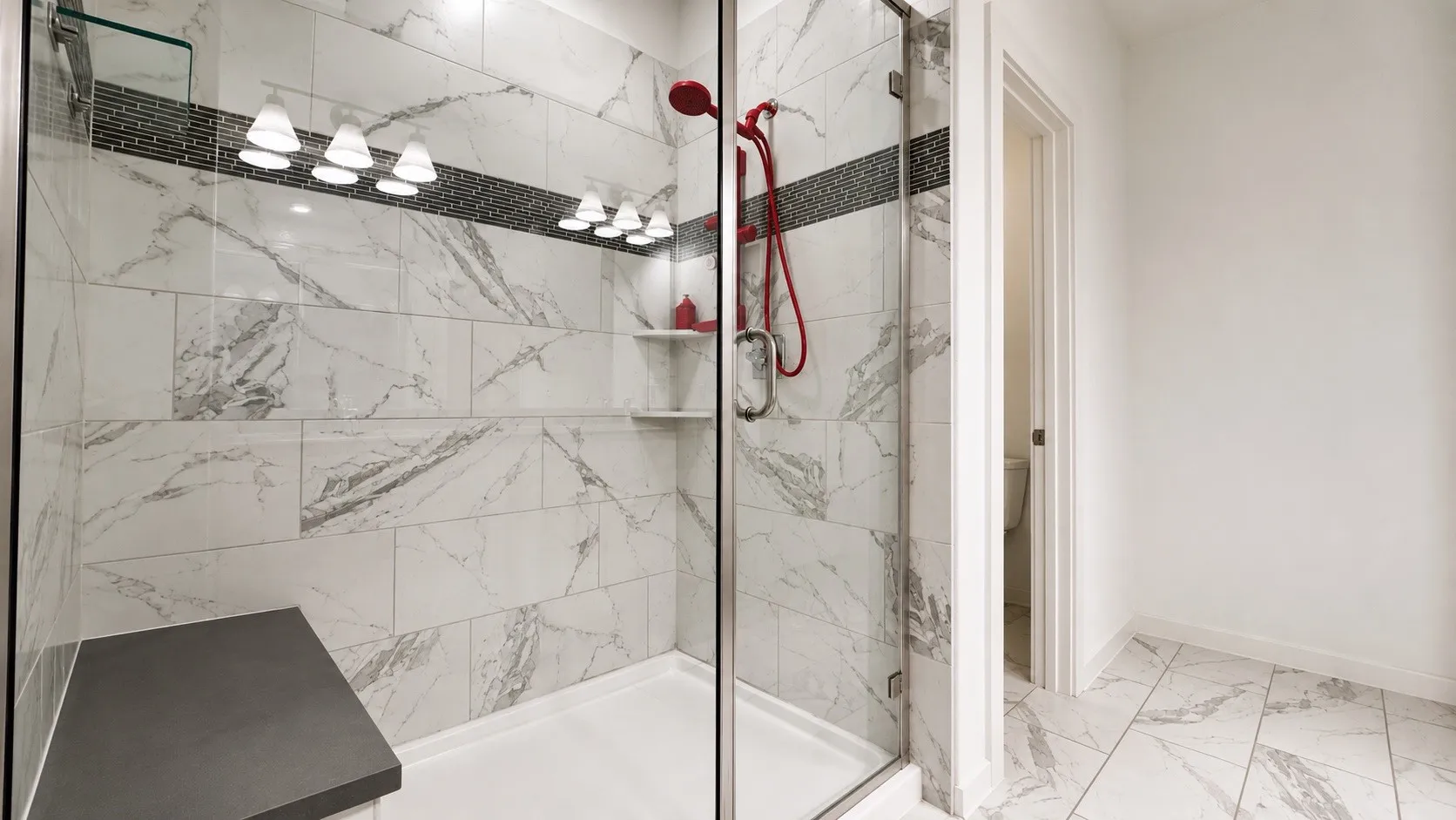Spacious shower enclosure featuring large format marble-look tile, a decorative mosaic accent band, and integrated shelving